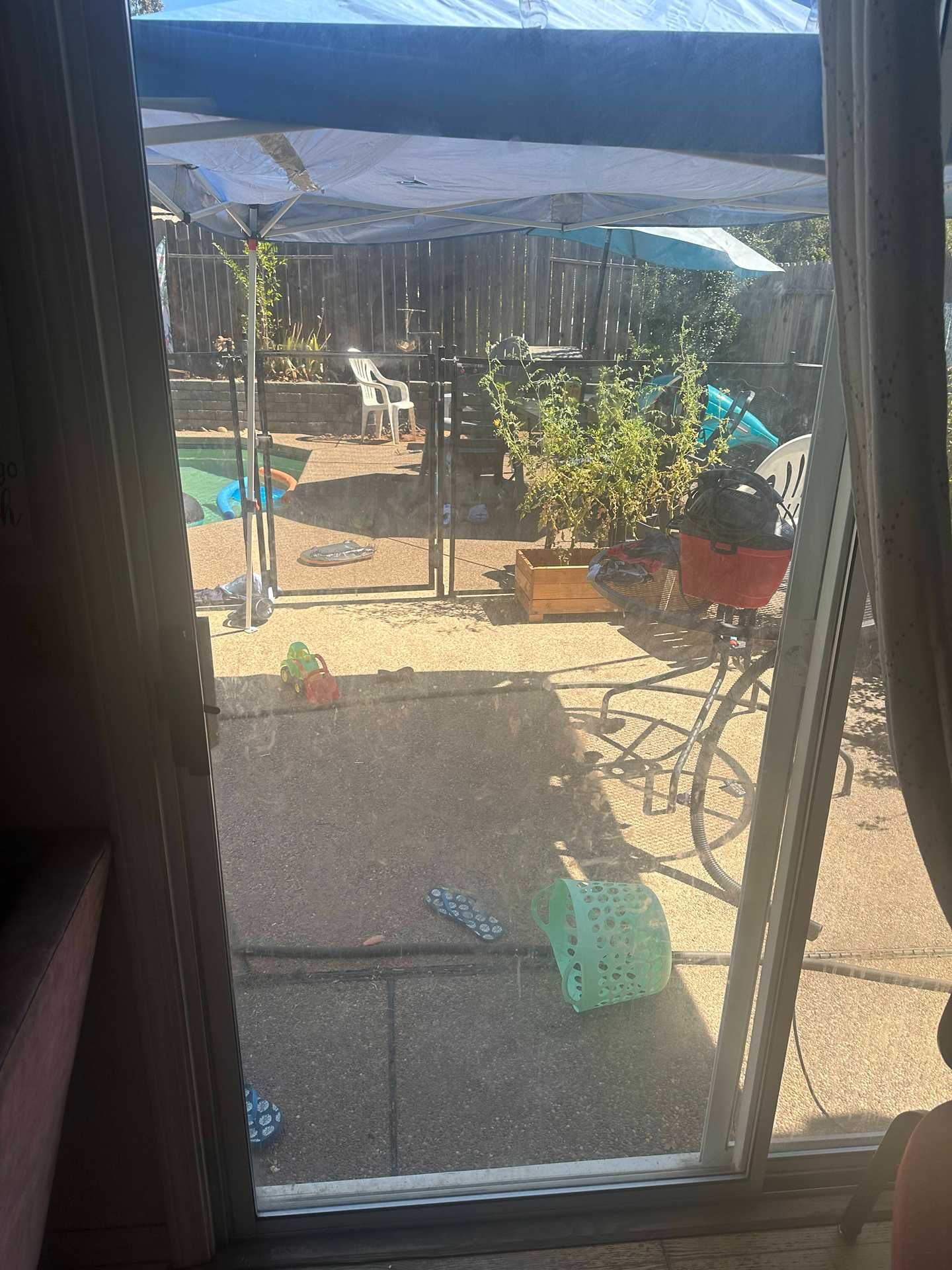 Patio seen through a window, with chairs, potted plants, and a blue tarp in bright sunlight.