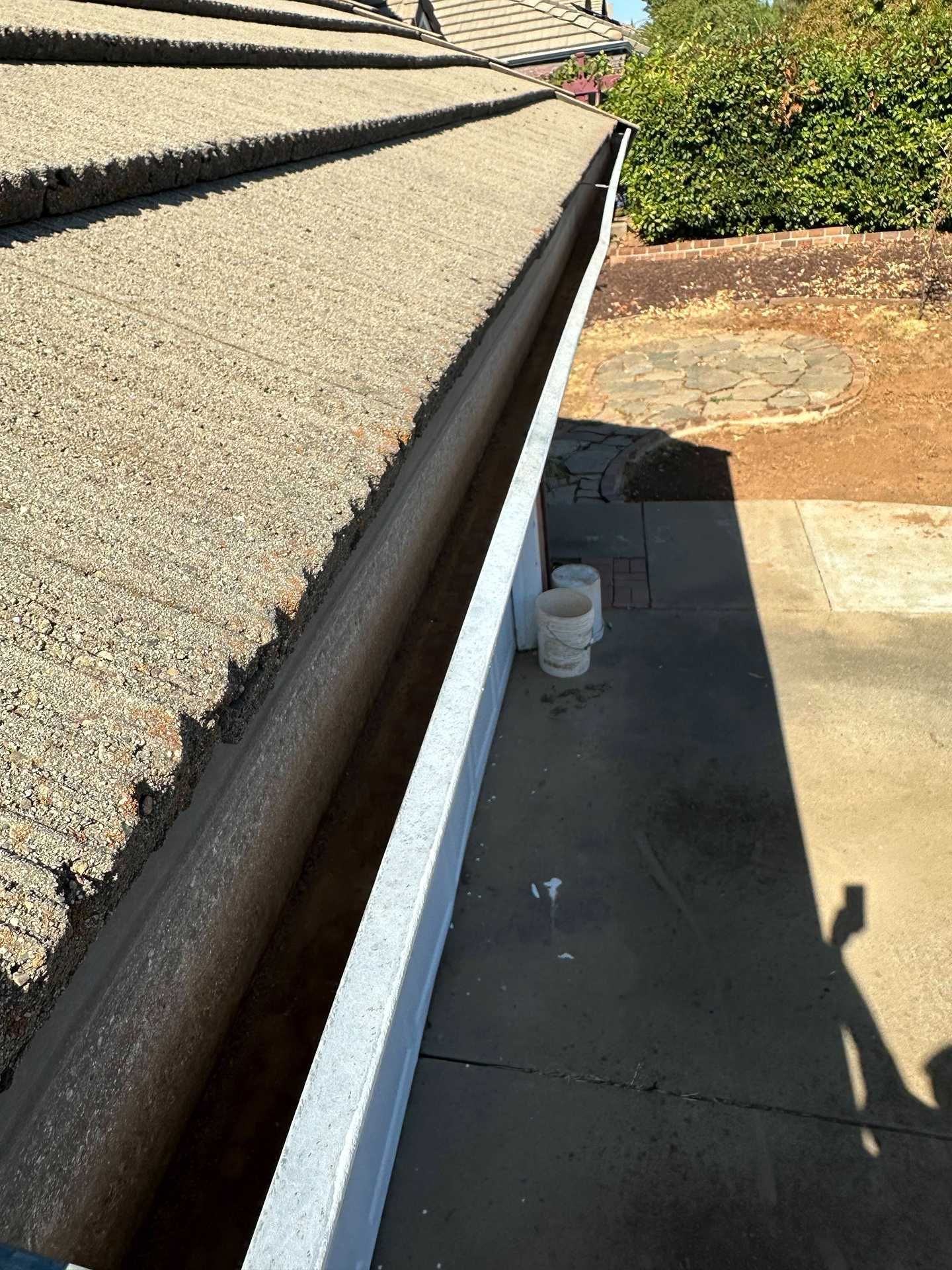 Close-up of roof eaves and gutter with white trim beside a concrete walkway and shrubbery.