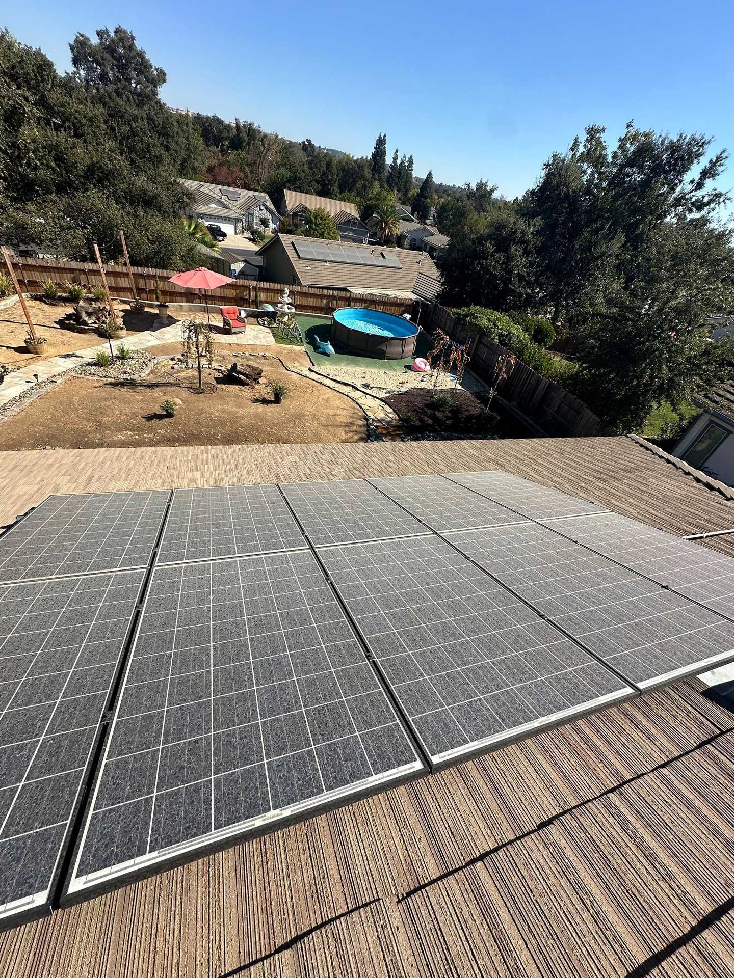 Solar panels on a roof overlooking a backyard with a blue pool and trees in bright sunlight.