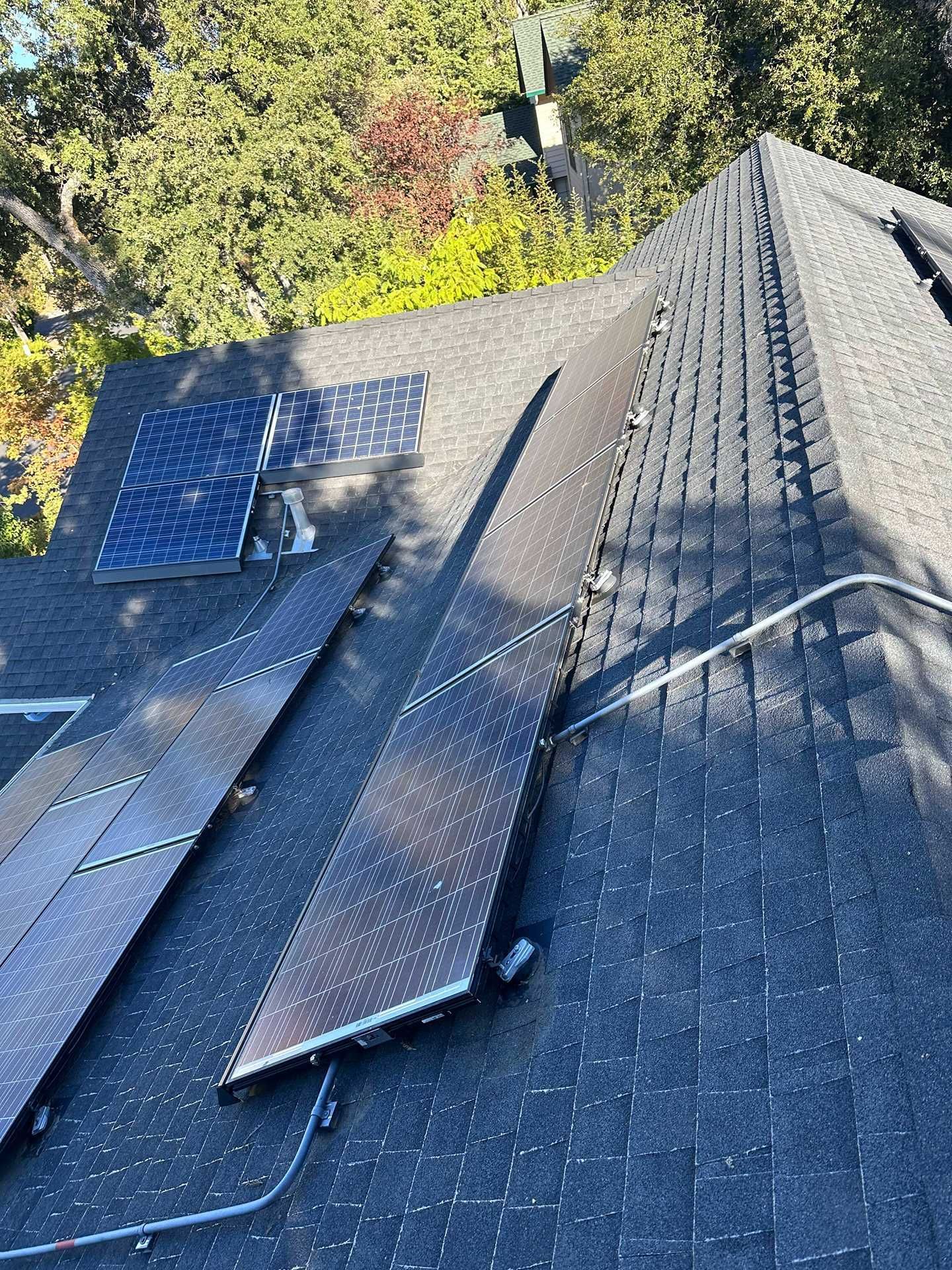 Solar panels and black roofing materials laid on a sloped roof beside trees.