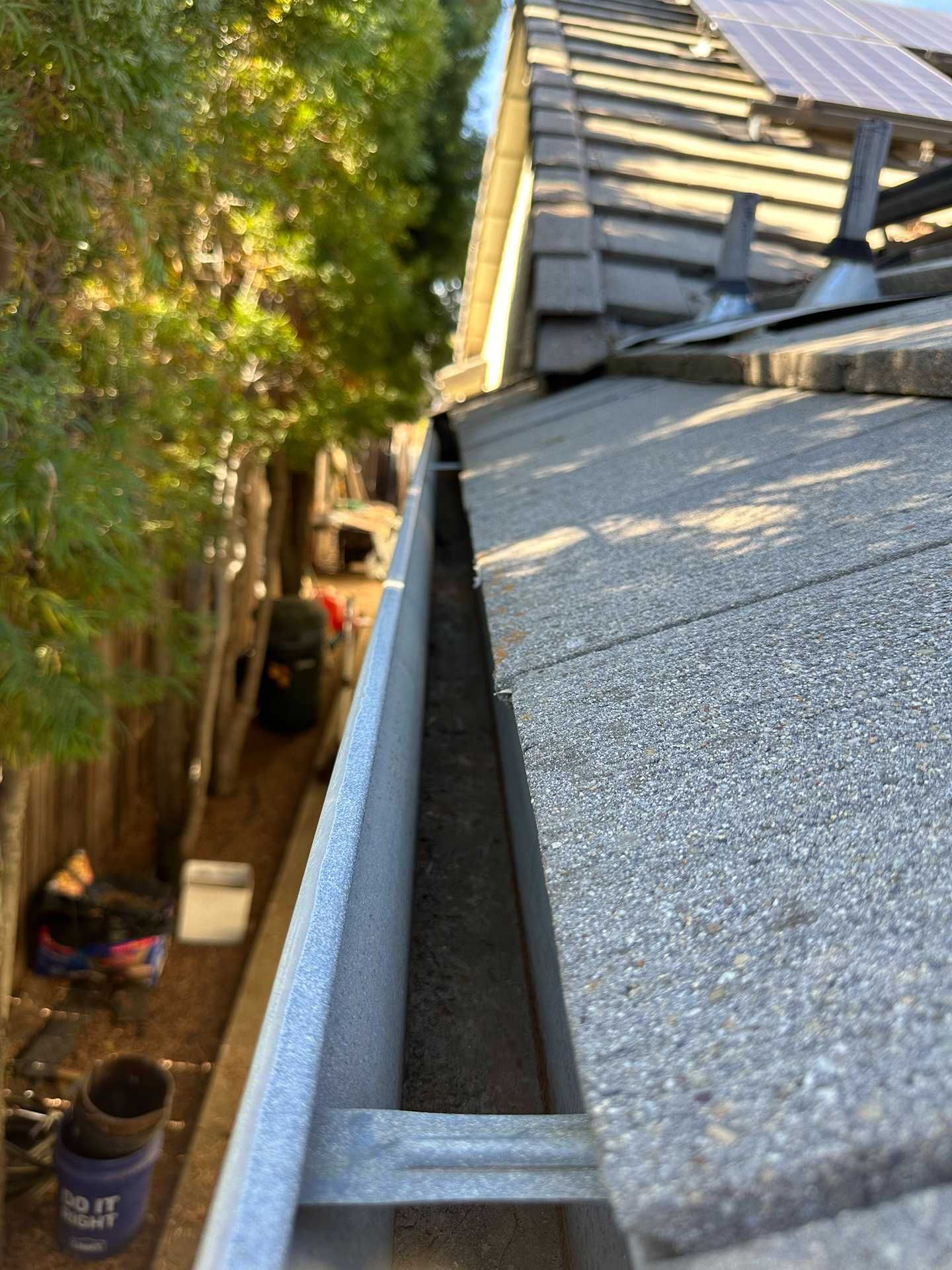 Close-up of a roof gutter beside a gray shingle roof, with trees and yard below