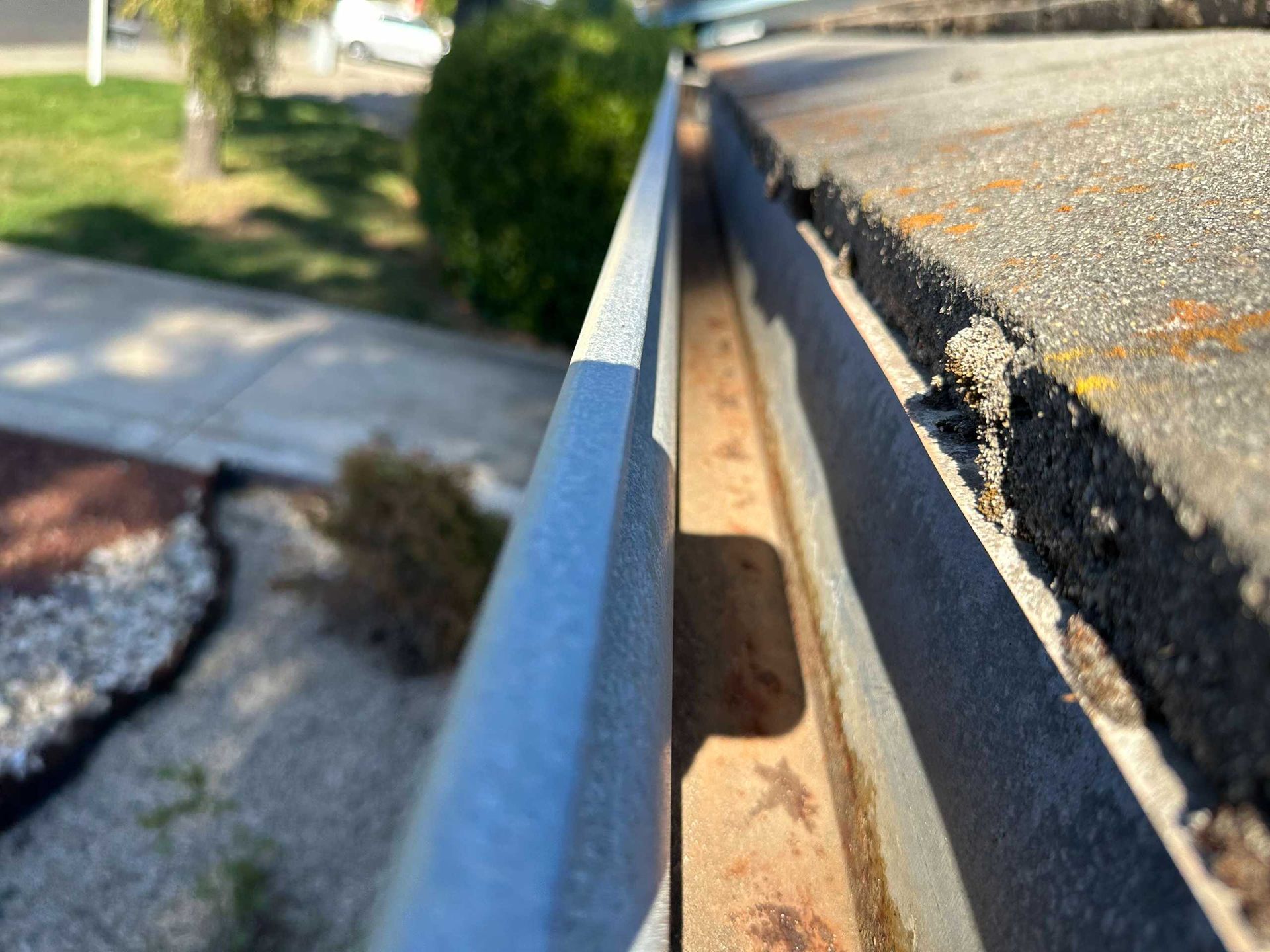 Close-up of a damaged roof gutter with peeling asphalt and rust beside a sidewalk