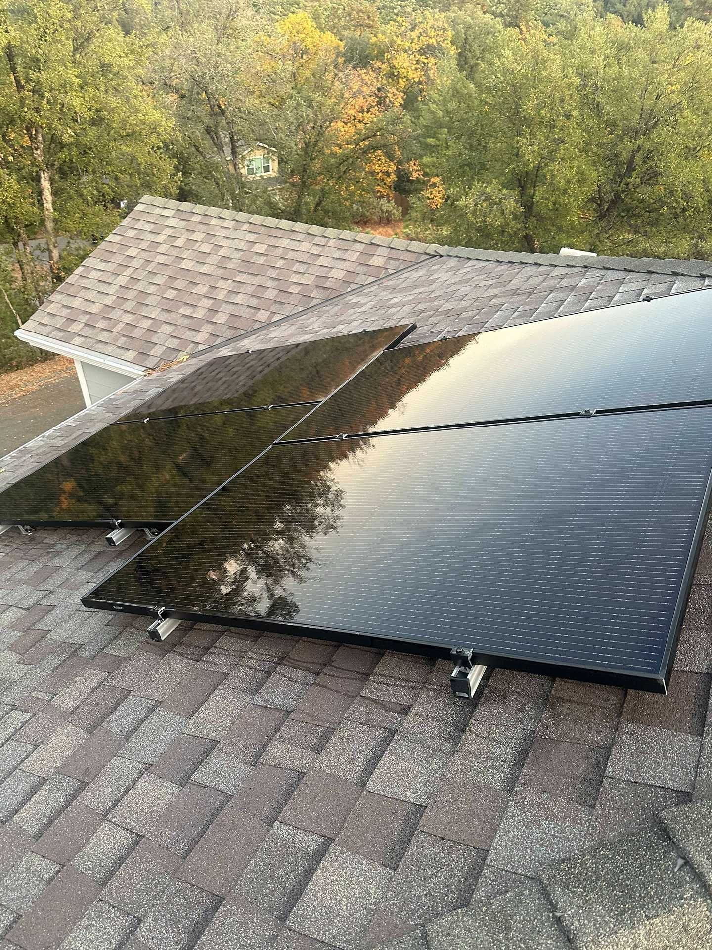 Solar panels mounted on a shingled roof beside a tree-lined yard at sunset