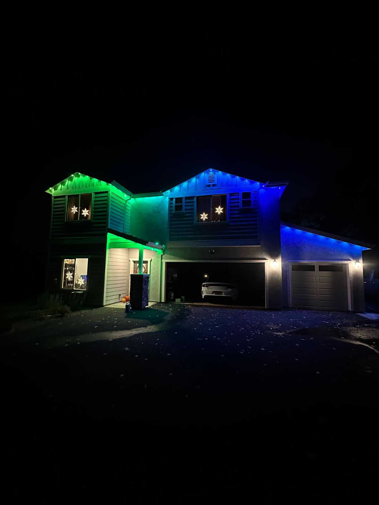 Two-story house at night with green and blue holiday lights and a lit garage driveway