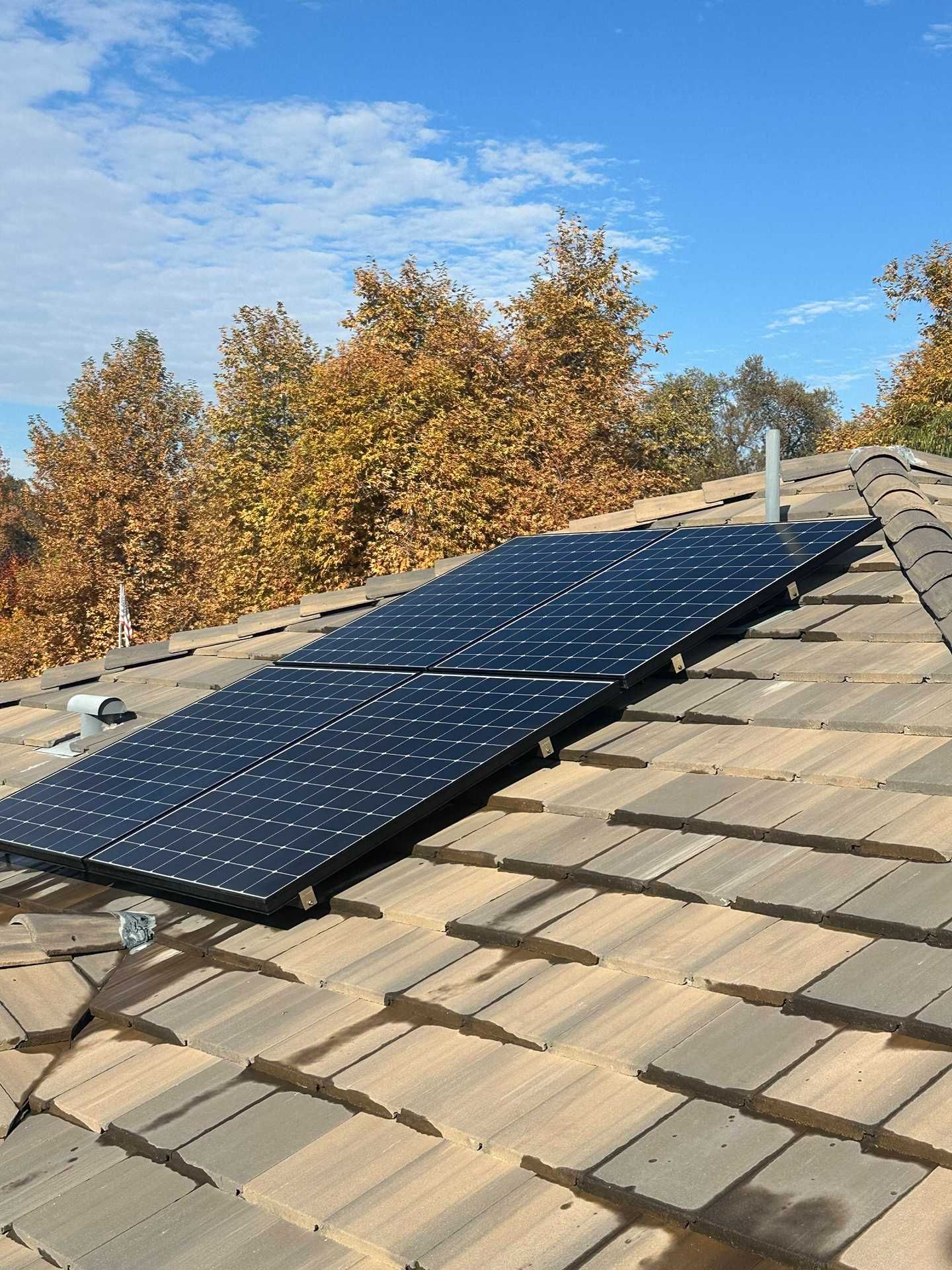 Solar panels mounted on a shingled roof under a blue sky, with autumn trees in the background