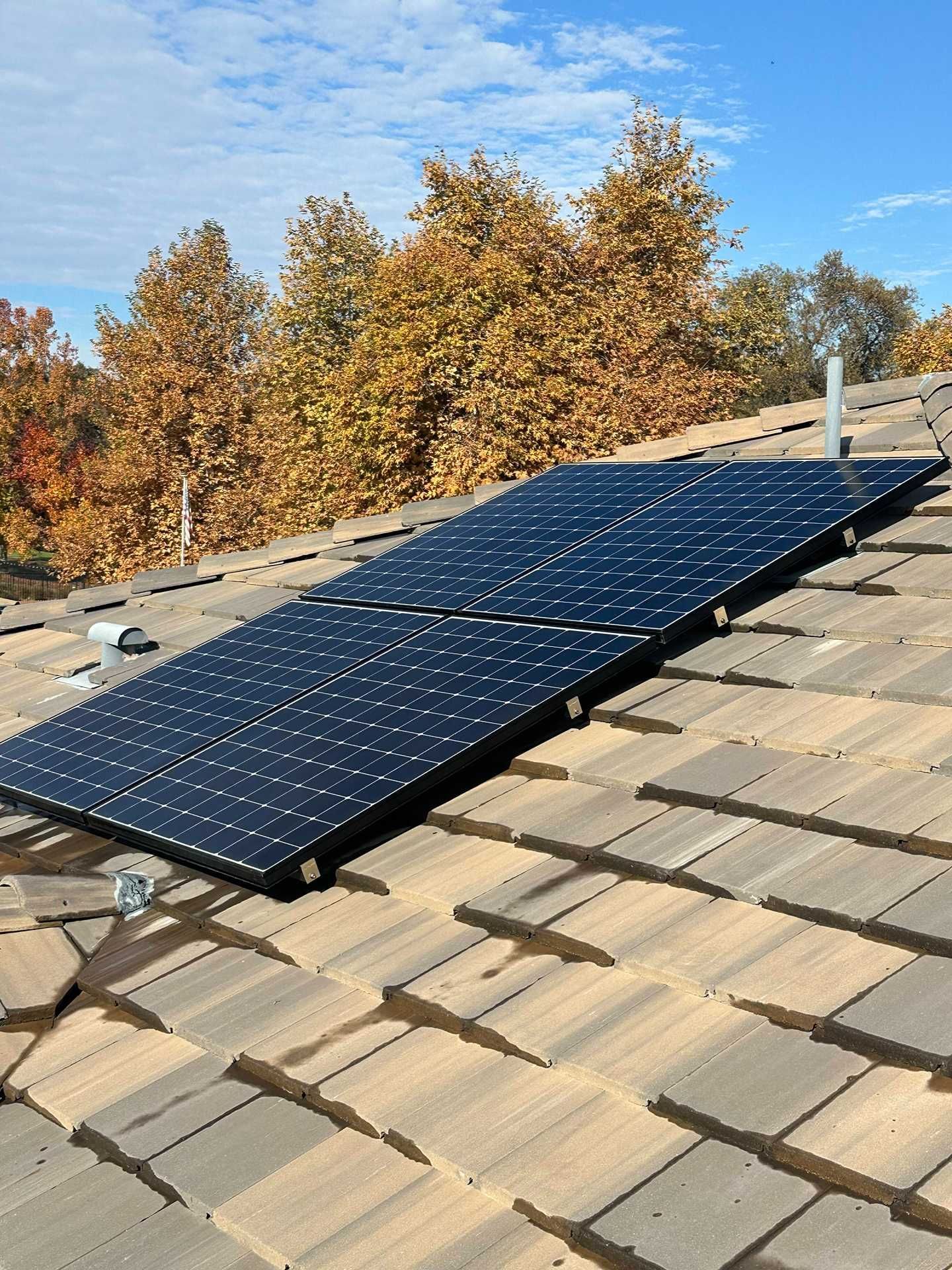 Solar panels installed on a tan tiled roof under a clear blue sky, with autumn trees in the background.