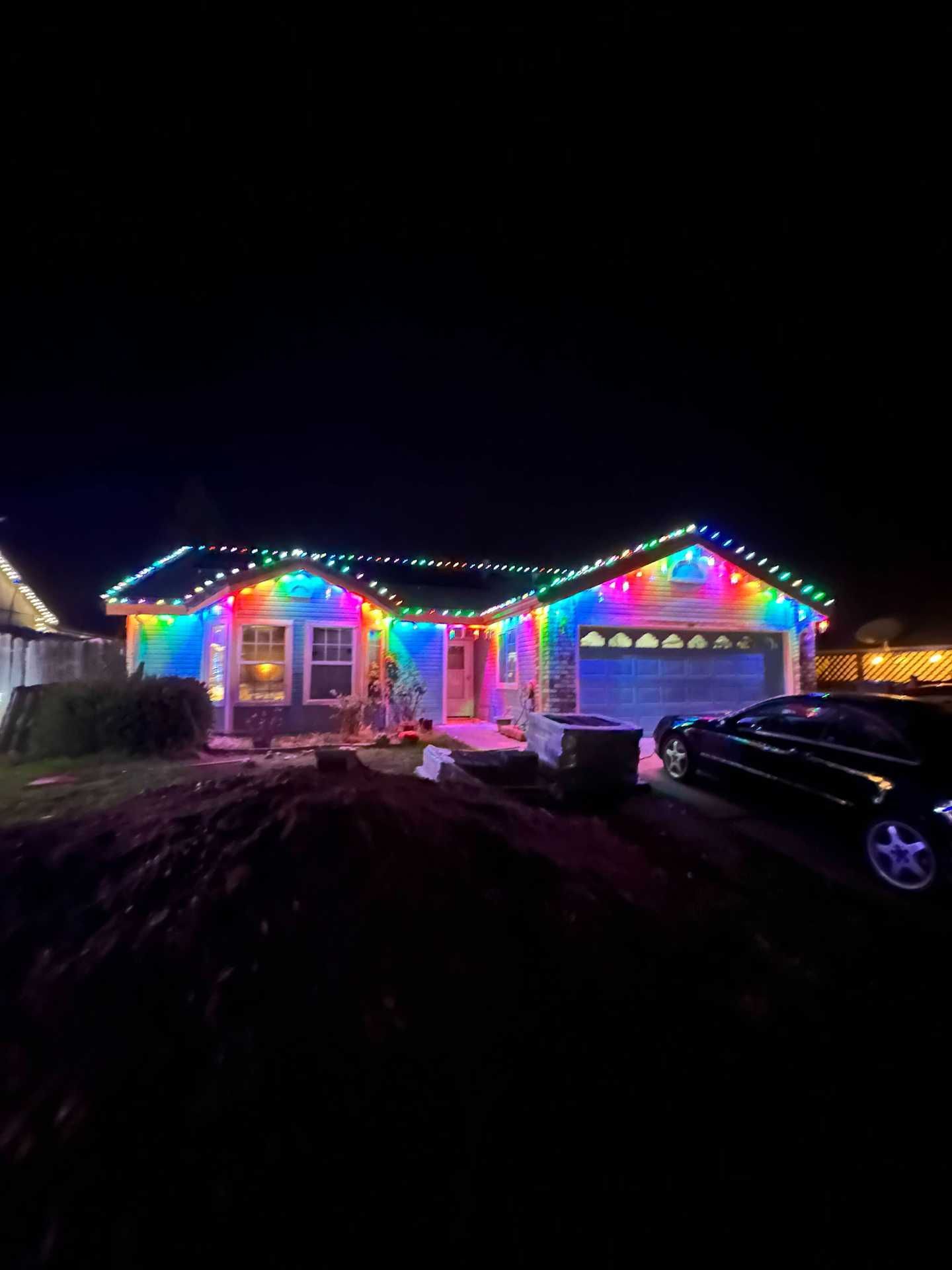 Single-story house outlined in rainbow holiday lights at night, with a parked car in the driveway.