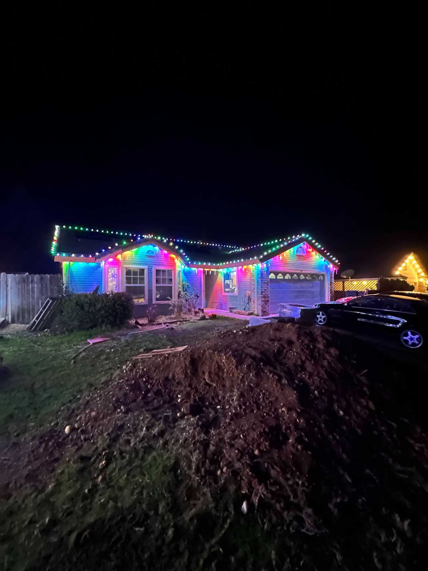 House with colorful Christmas lights at night, with a mound of dirt in the foreground and a parked car on the right