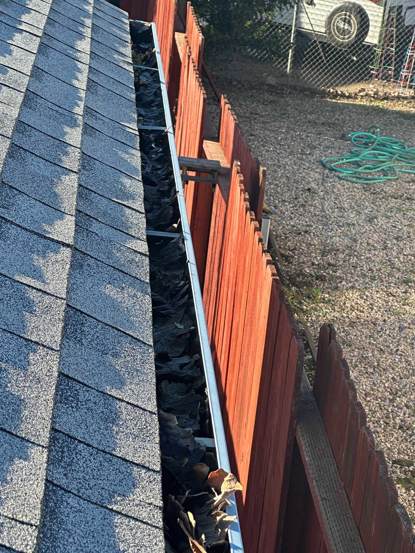 Roof shingles beside a narrow gutter over a backyard fence and gravel yard