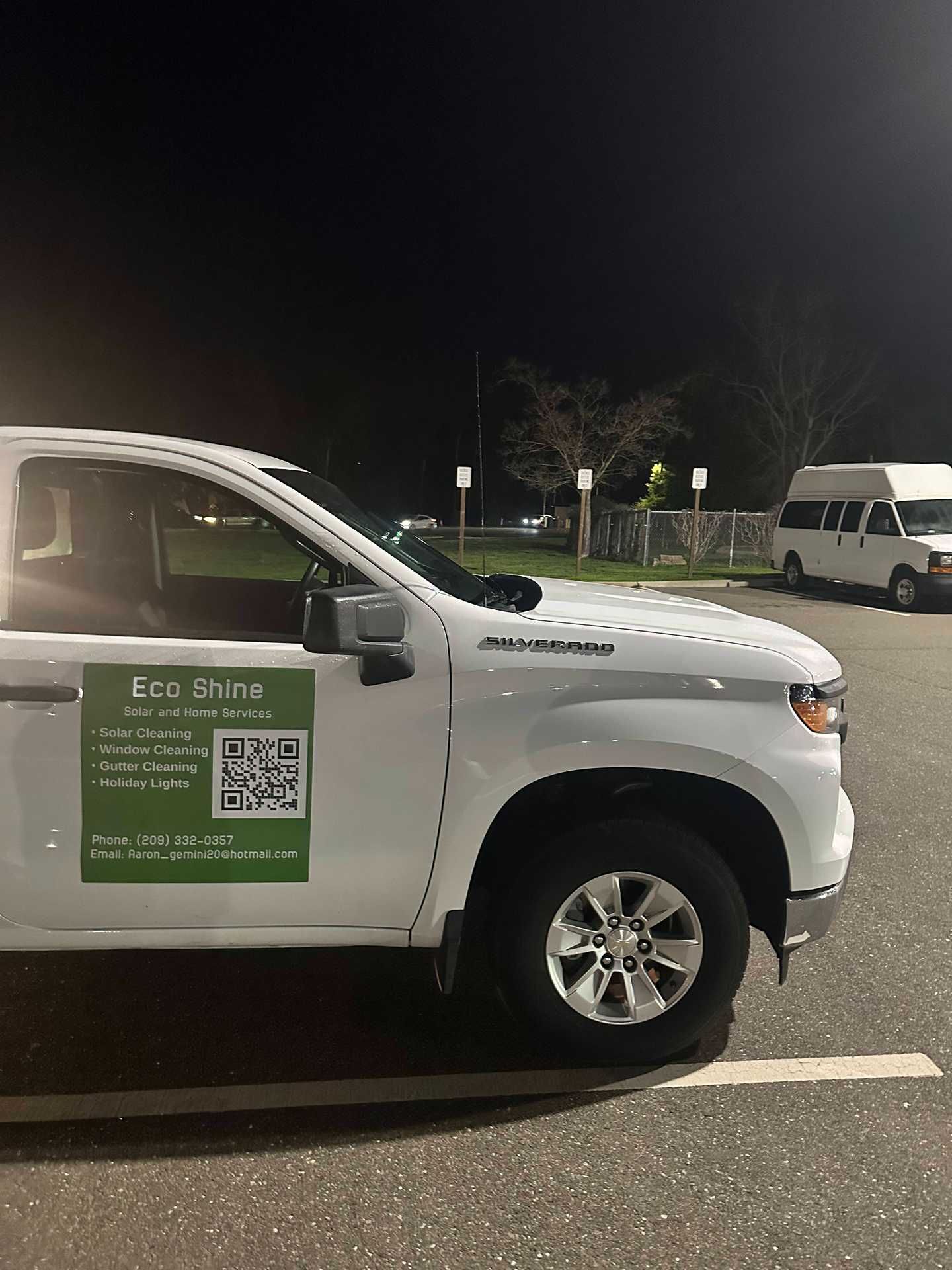 White pickup with a green QR code sign parked at night in a lot, with a van in the background.