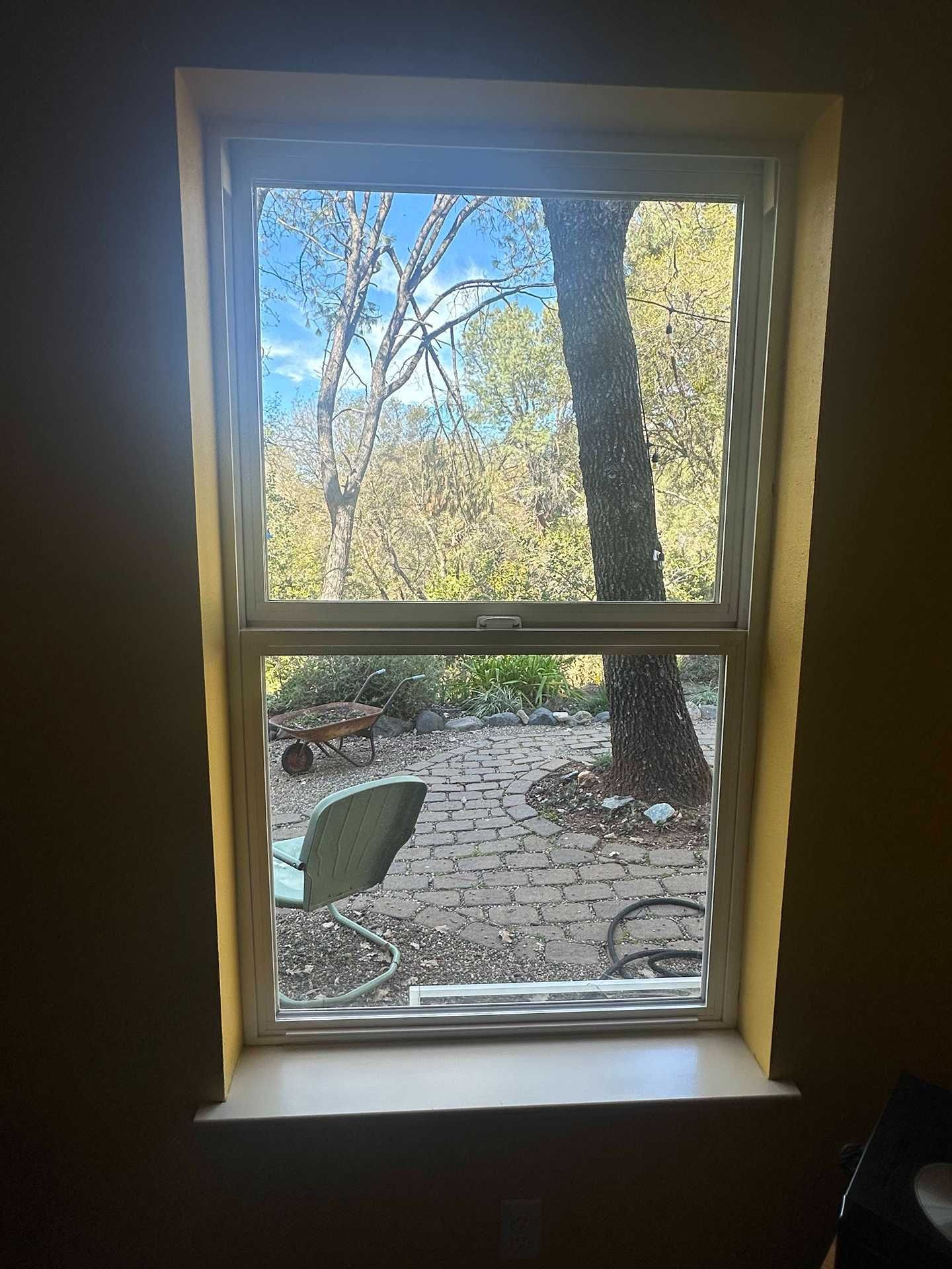 View through a window to a stone patio with a chair, trees, and bright daylight beyond.
