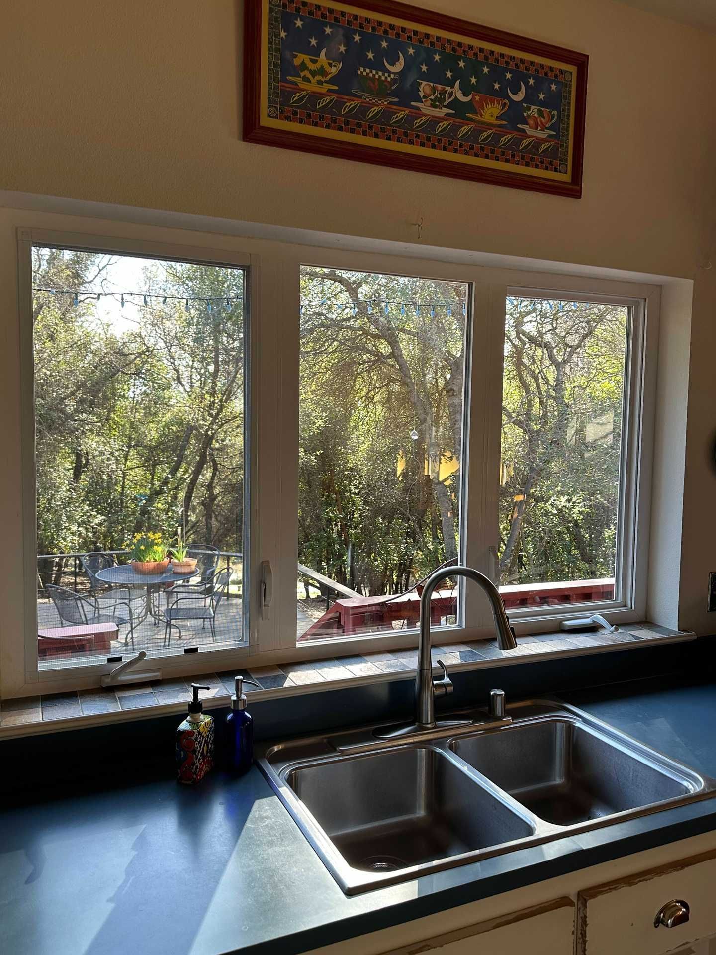 Kitchen sink beneath a window overlooking trees, with blue countertop and red-framed wall art above.