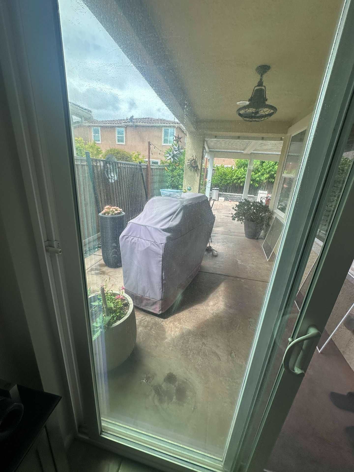 Covered patio viewed through a glass door, with a covered grill, potted plants, and ceiling fan.