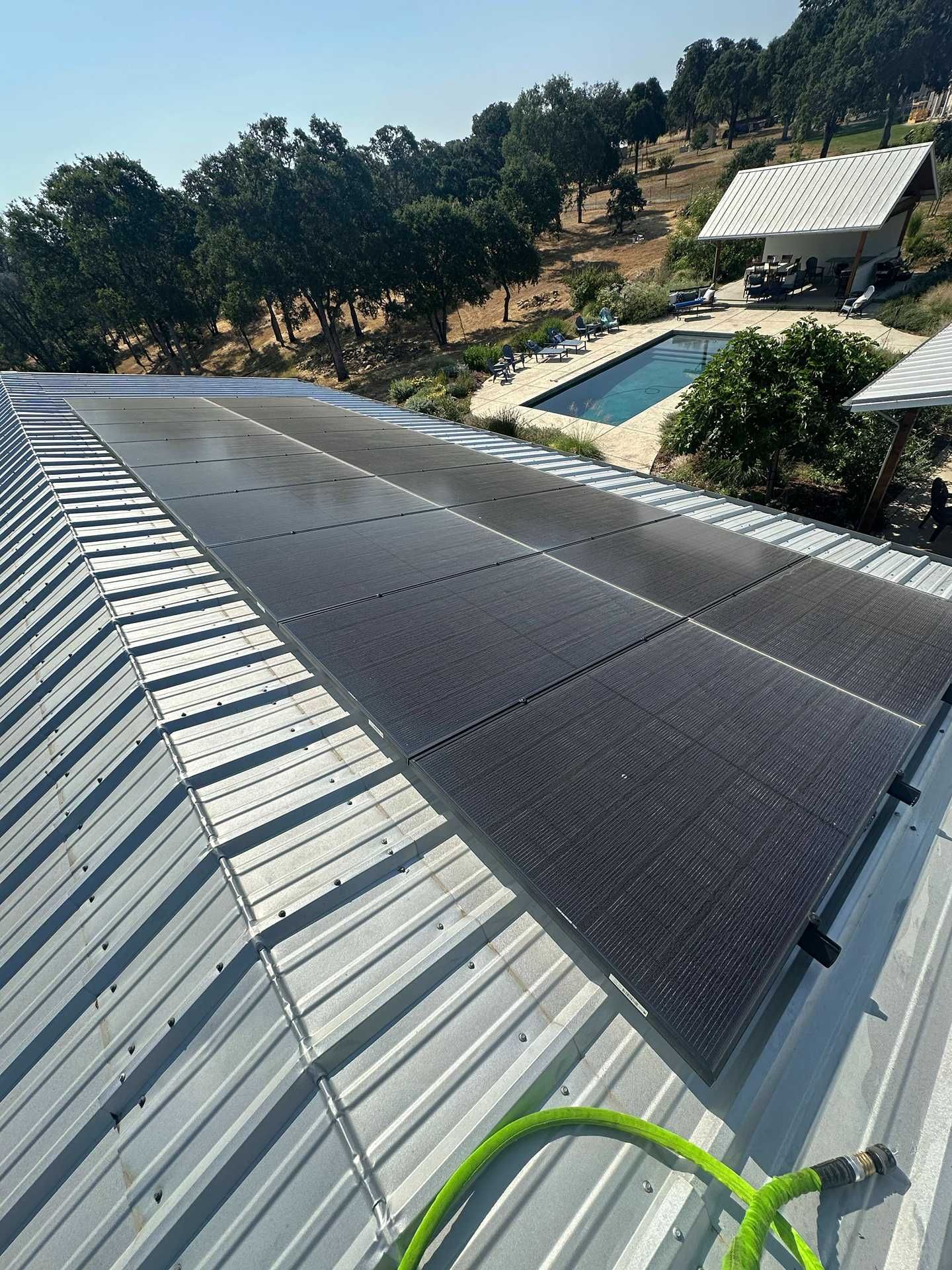 Solar panels on a metal roof beside a pool and hillside trees, with a green hose in the foreground