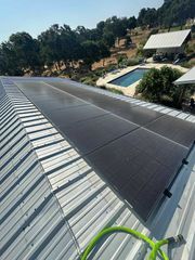 Solar roof panels on a sloped metal roof beside a pool and hillside trees.