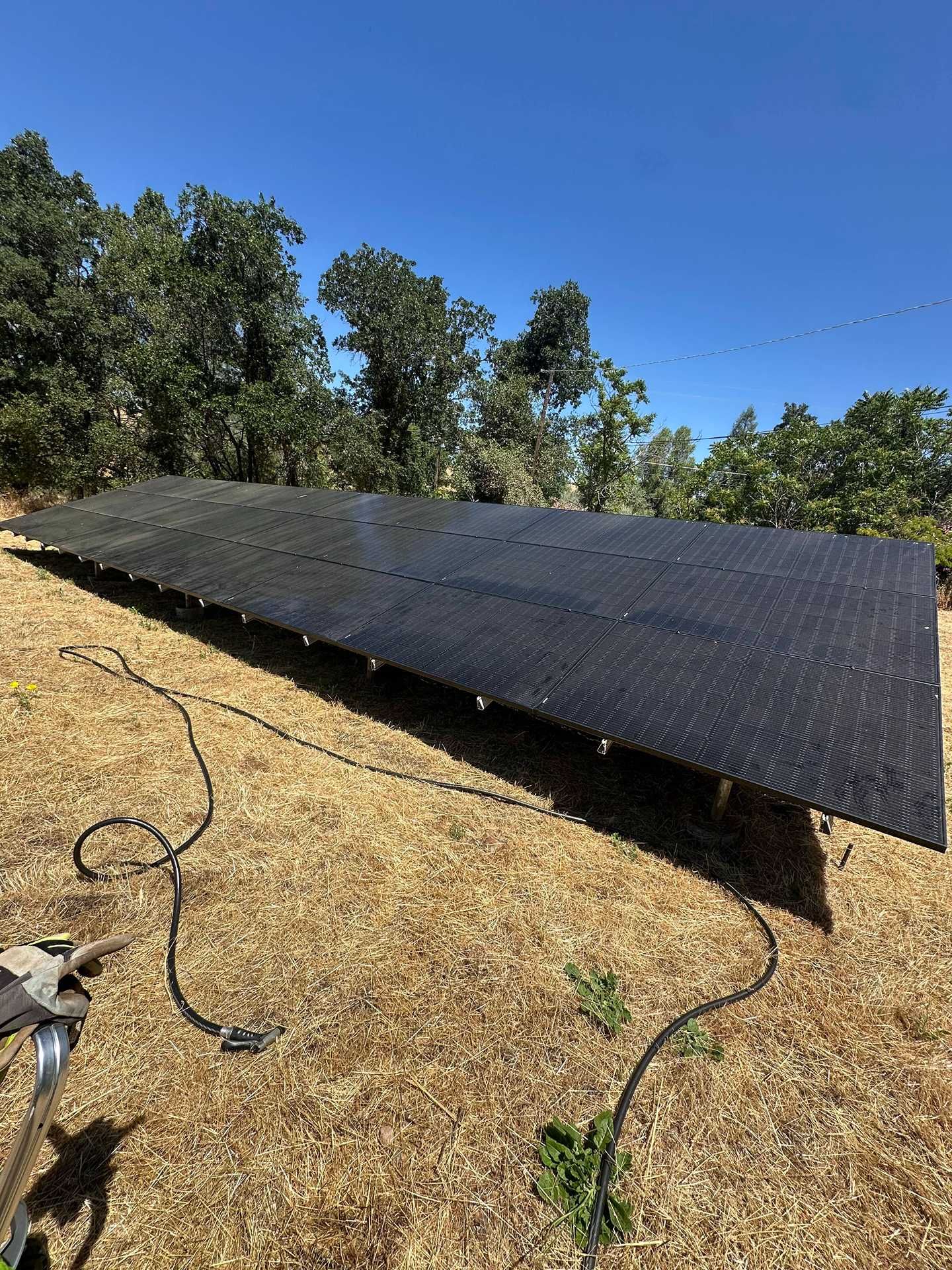 Black solar panel array on dry hillside, with trees and blue sky in the background.