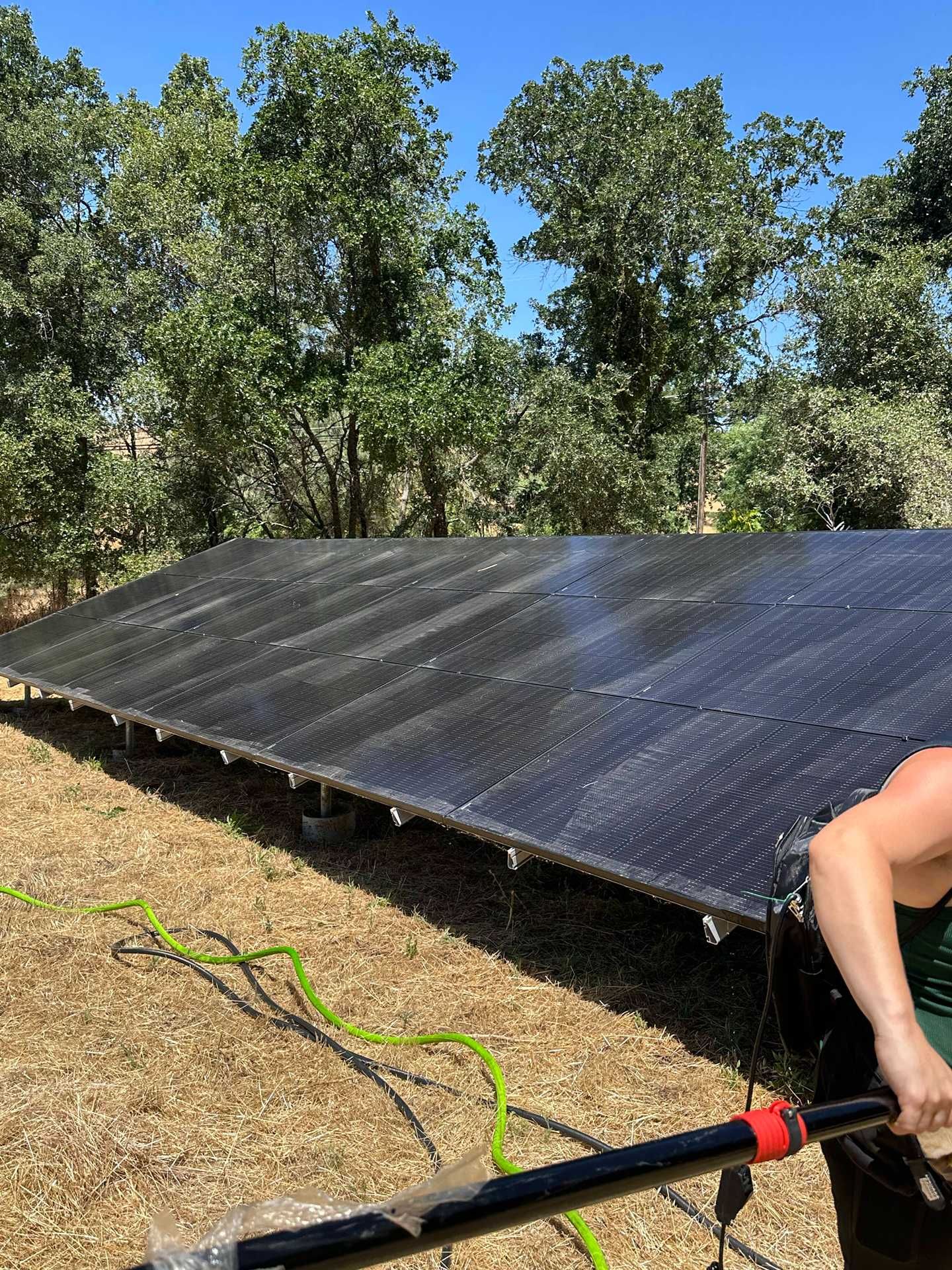Solar panels installed on a dry hillside, with trees in the background and a worker partially visible at right.