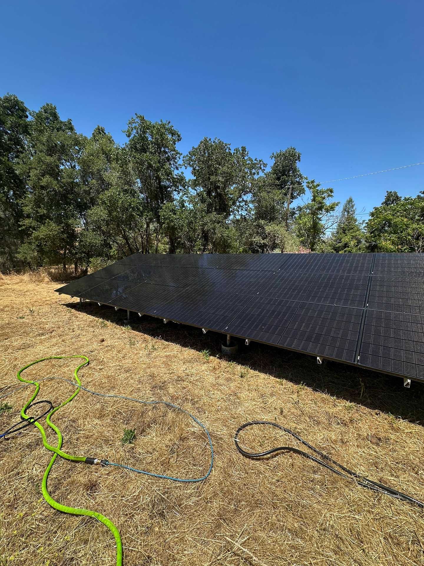 Row of black solar panels on dry ground in a sunny outdoor field with green hose in foreground