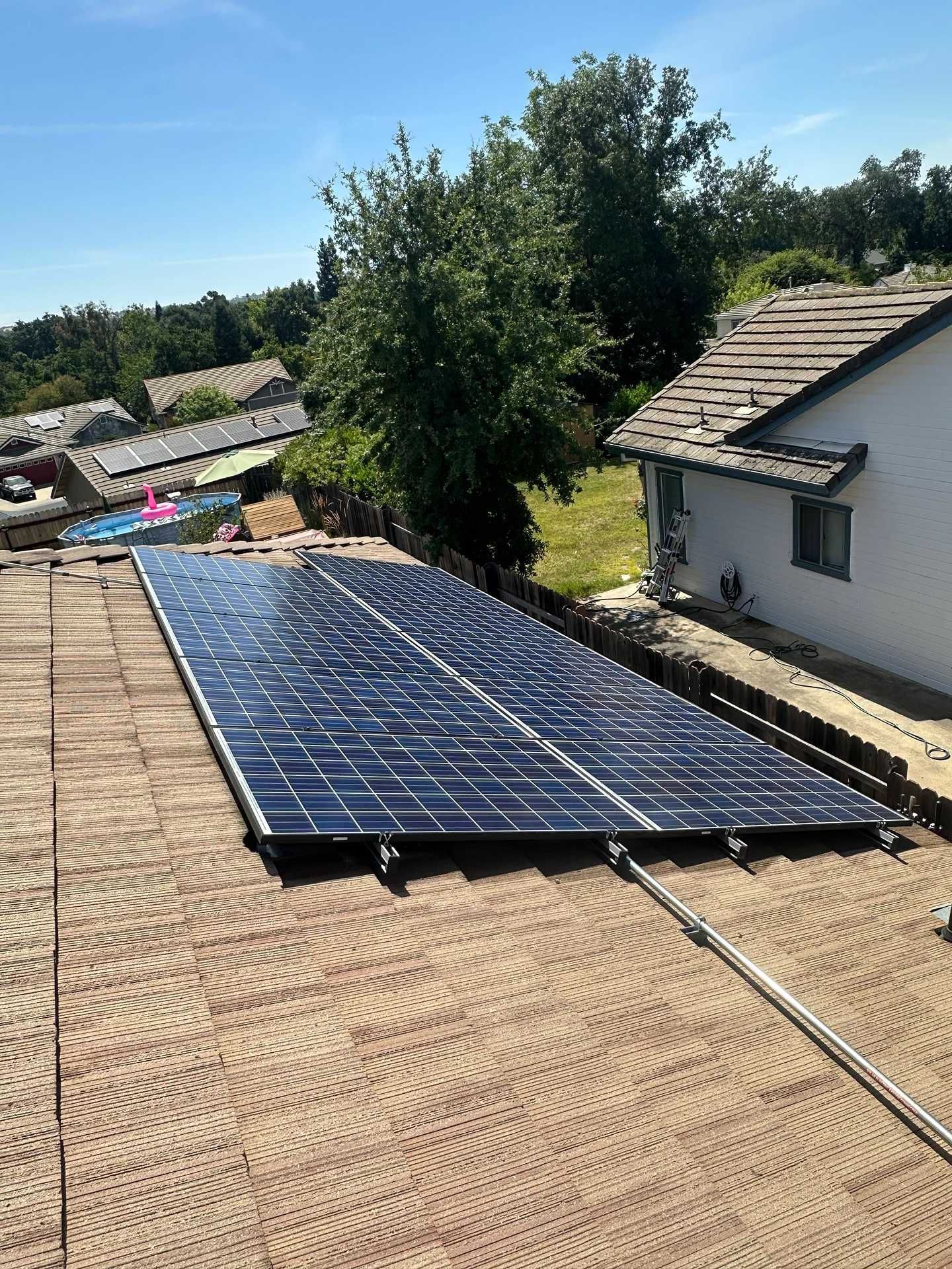 Solar panels installed on a tan shingle roof beside a white house and trees.