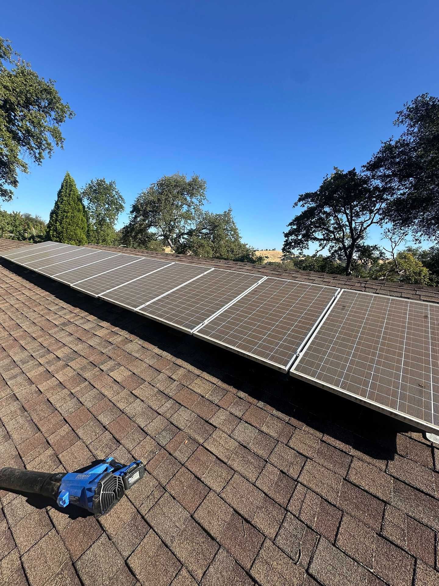 Solar panels installed across a shingled roof under a clear blue sky, with trees nearby.