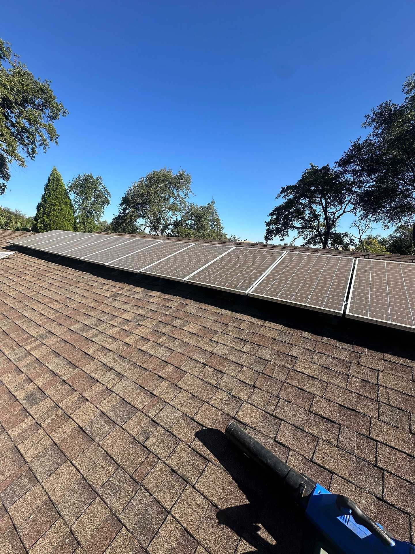 Roof with solar panels under a clear blue sky, viewed over brown shingles from a rooftop edge.