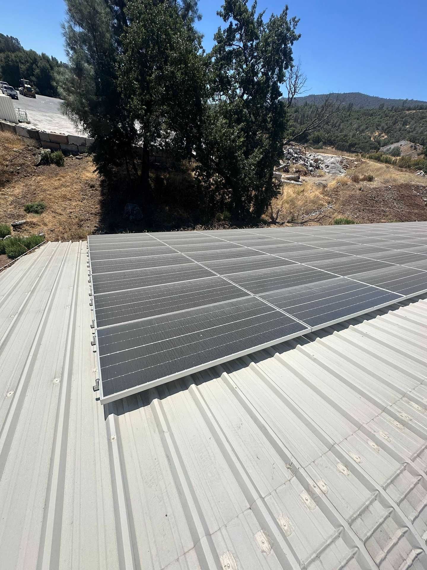 Solar panels mounted on a light gray metal roof with hills and trees in the background