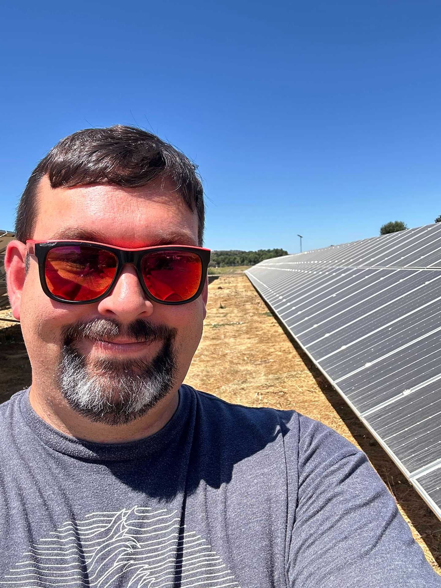Person wearing red sunglasses taking a selfie beside solar panels under a blue sky