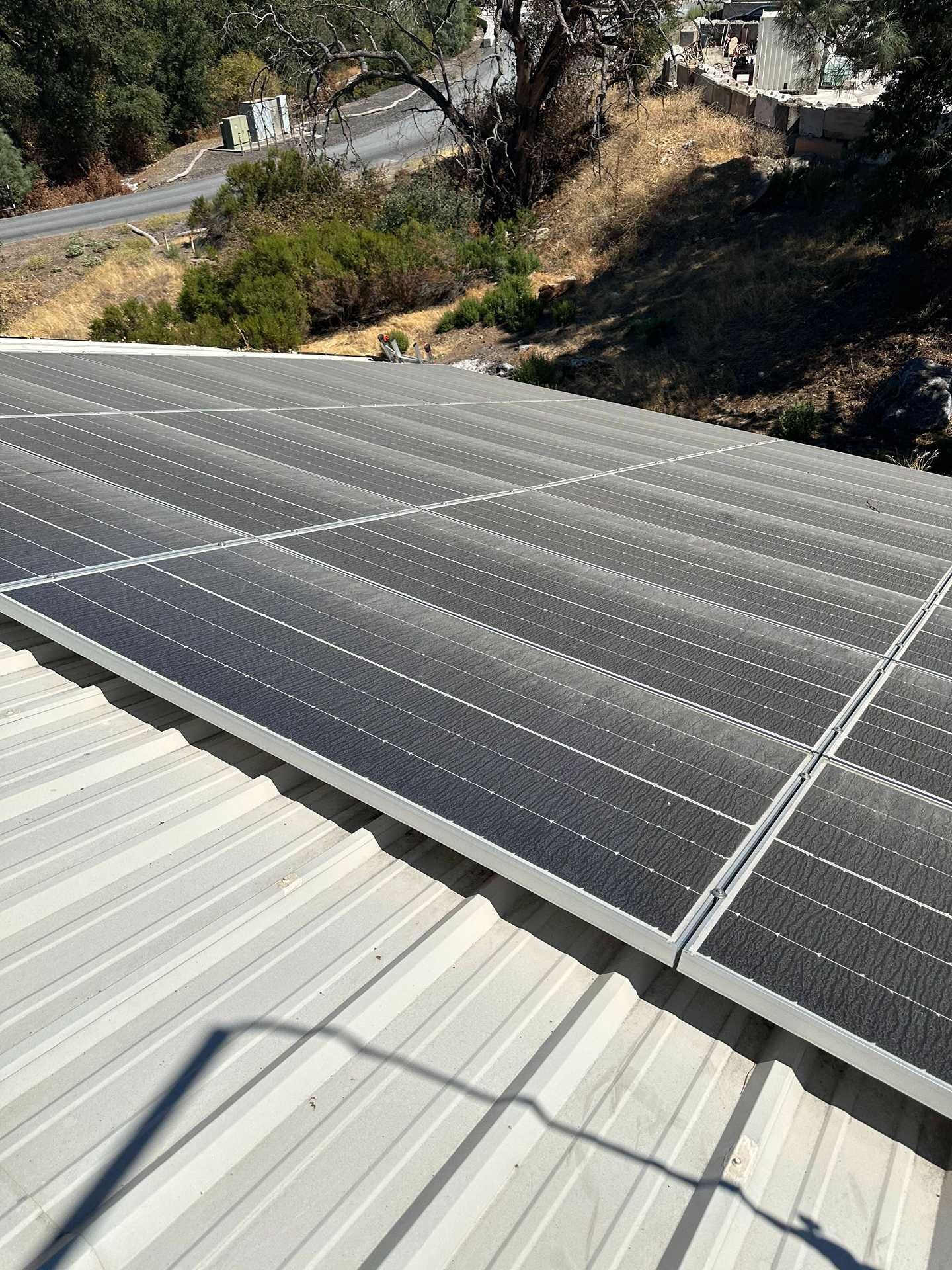 Solar panels on a white metal roof overlooking a dry hillside road and trees.