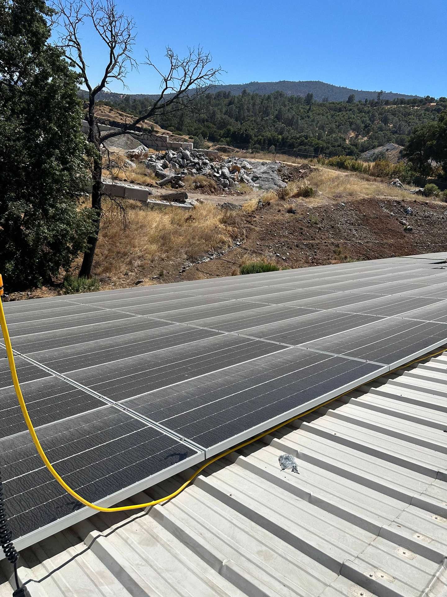 Solar panels on a rooftop overlooking dry hills and trees under a clear sky.