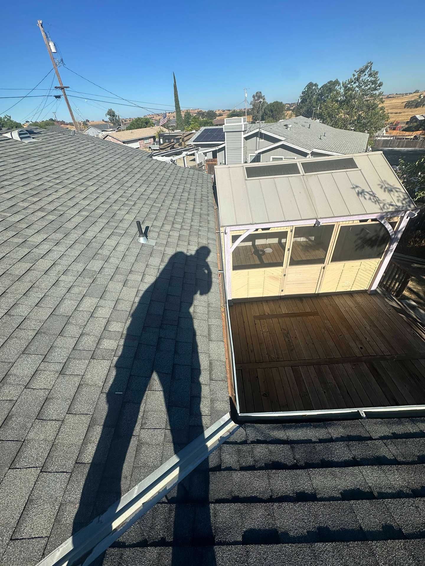 Roof view with gray shingles, rooftop shadow, and a white pergola over a wooden deck under blue sky.