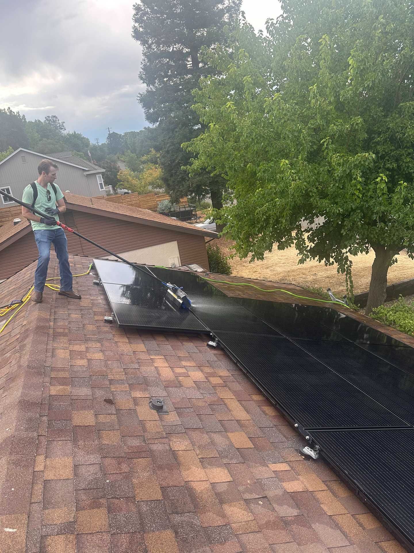 Worker inspecting a sloped roof with black underlayment and loose shingles beside trees and houses
