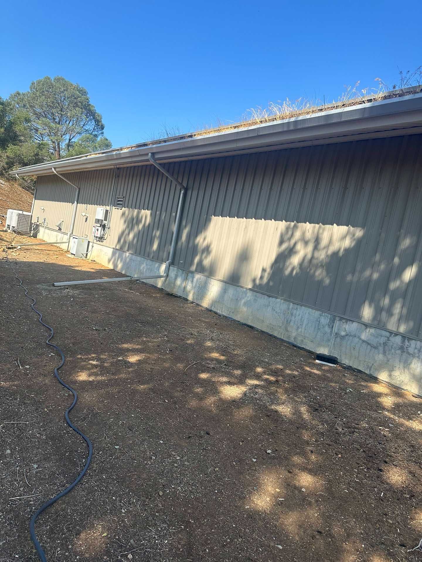 Long gray building with a sloped roof beside a dirt lot and trees in sunlight