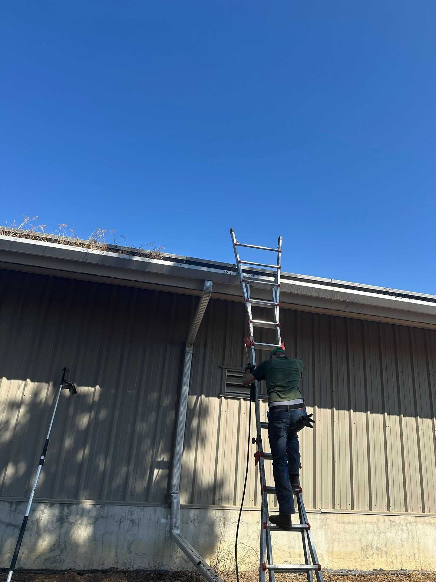 Worker on a ladder reaching toward a roof gutter beside a gray building under a clear blue sky