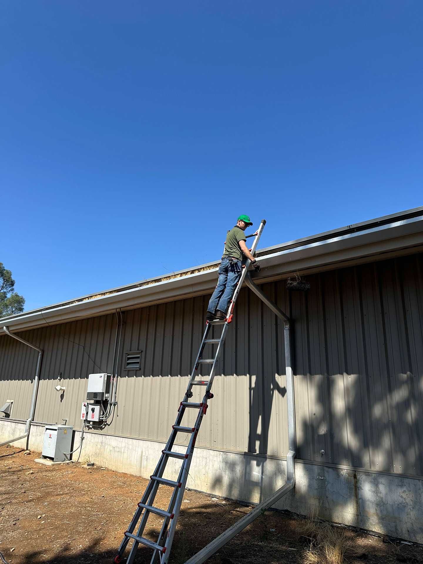Worker on a ladder repairing the roof edge of a beige building under a clear blue sky.