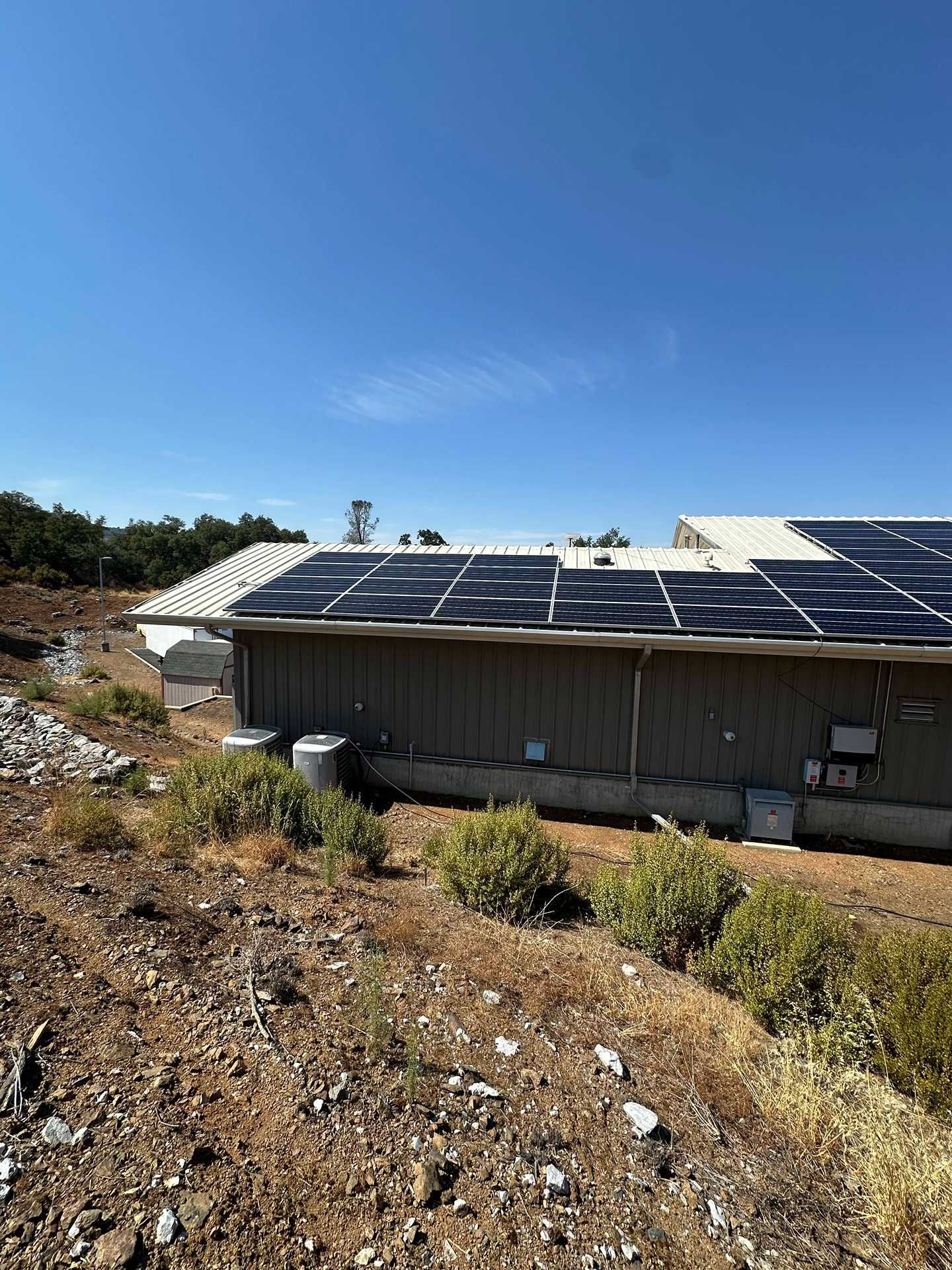 Rural building with solar panels on the roof under a clear blue sky, surrounded by dry brush.