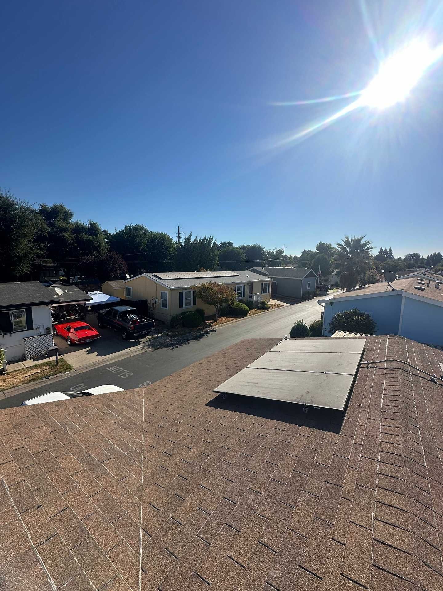 Sunlit suburban street with parked cars, houses, trees, and a roof in the foreground