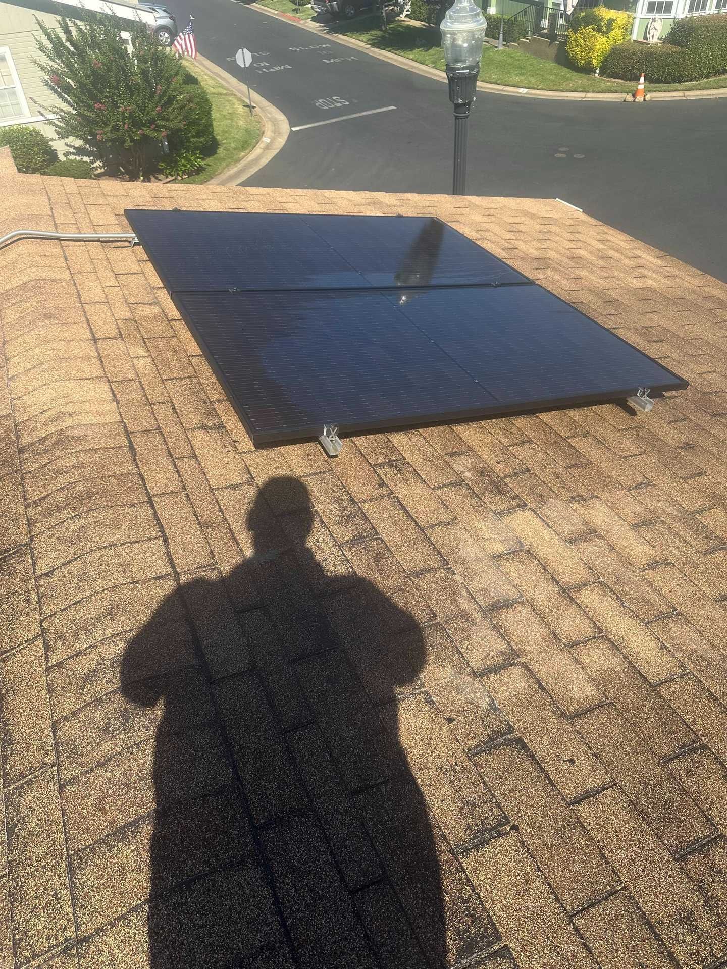 Black solar panel on a shingled rooftop, with a shadow in the foreground and a street in the background