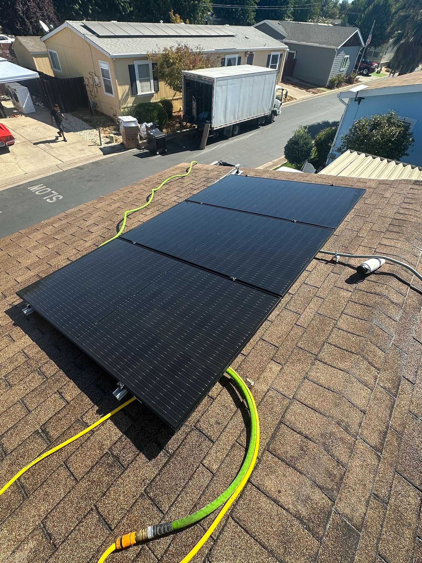 Solar panels on a shingled roof in a suburban neighborhood, with yellow and green hoses nearby.