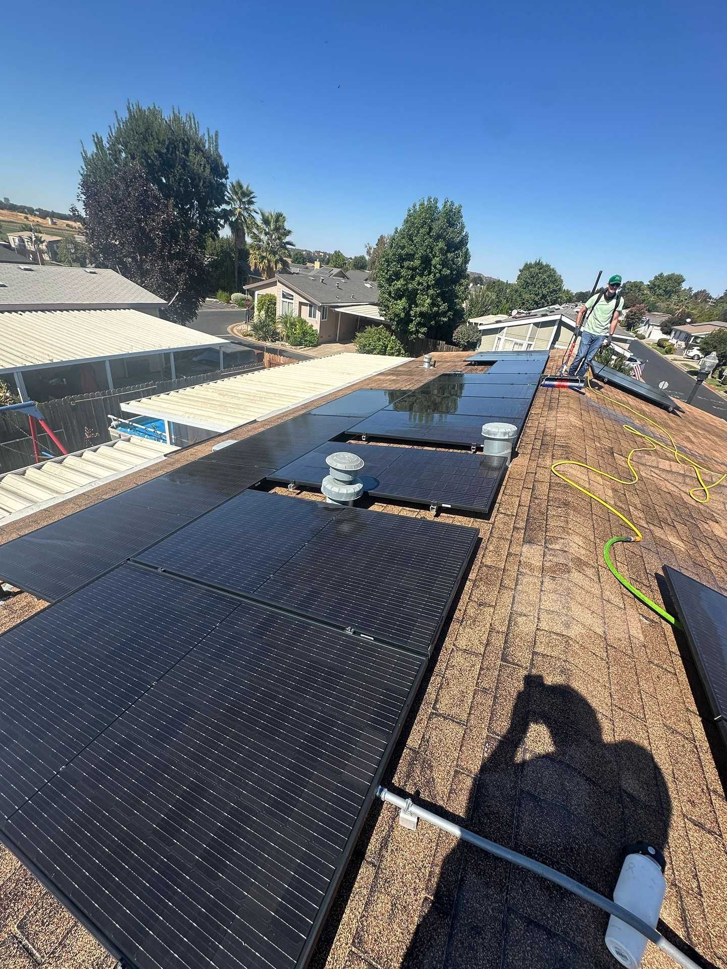 Solar panels installed on a rooftop under a sunny sky, with trees and neighboring houses in the background.