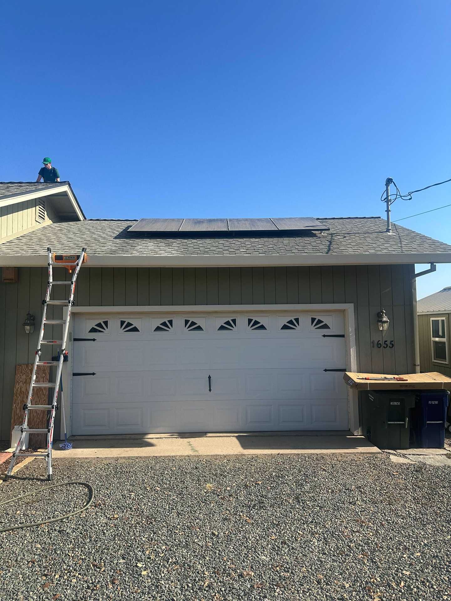 Single-story house with white garage door, ladder on left roof, and trash bins on gravel driveway.