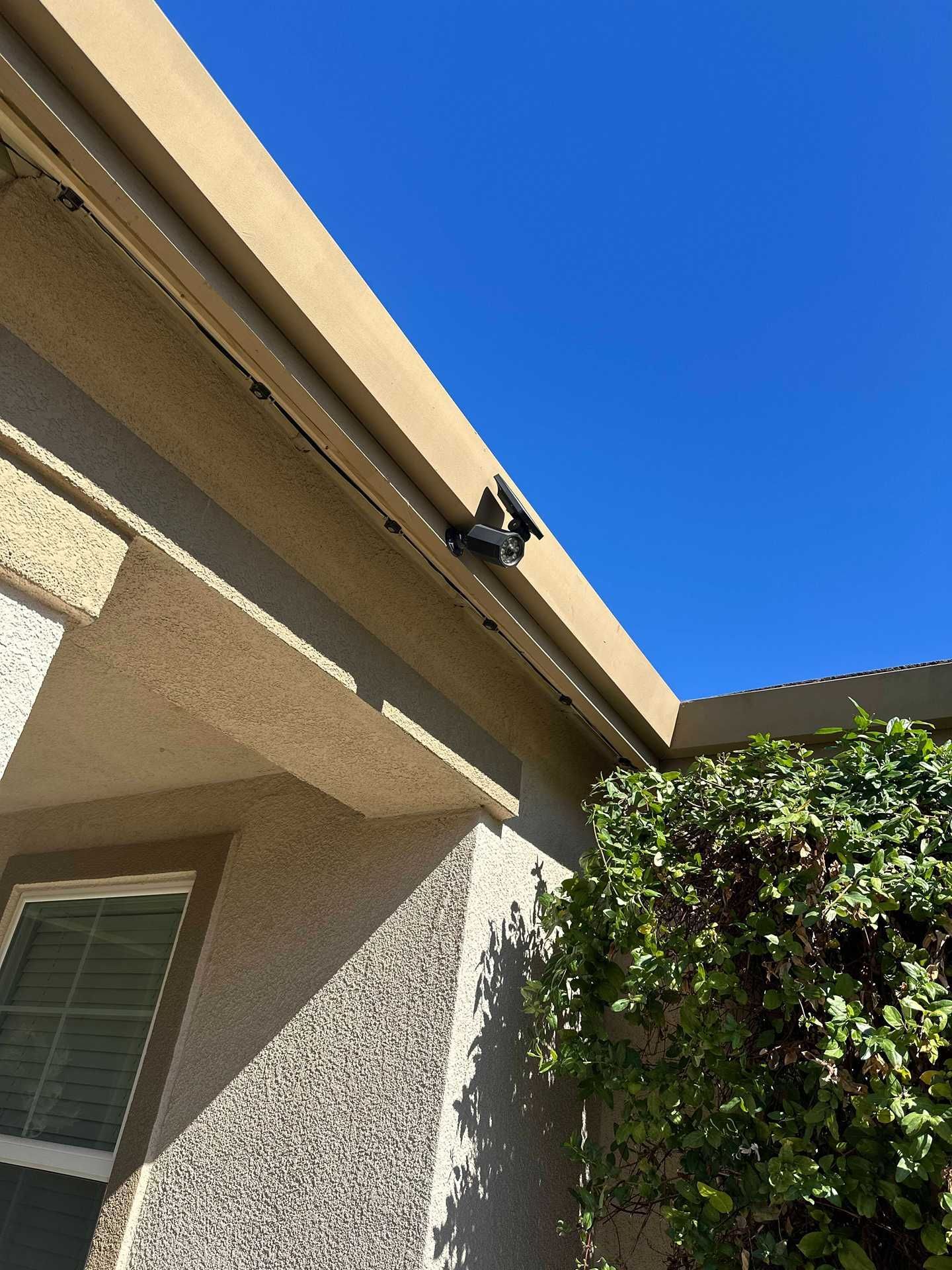 Corner of a house roof and stucco wall against a blue sky, with a security camera under the eaves.