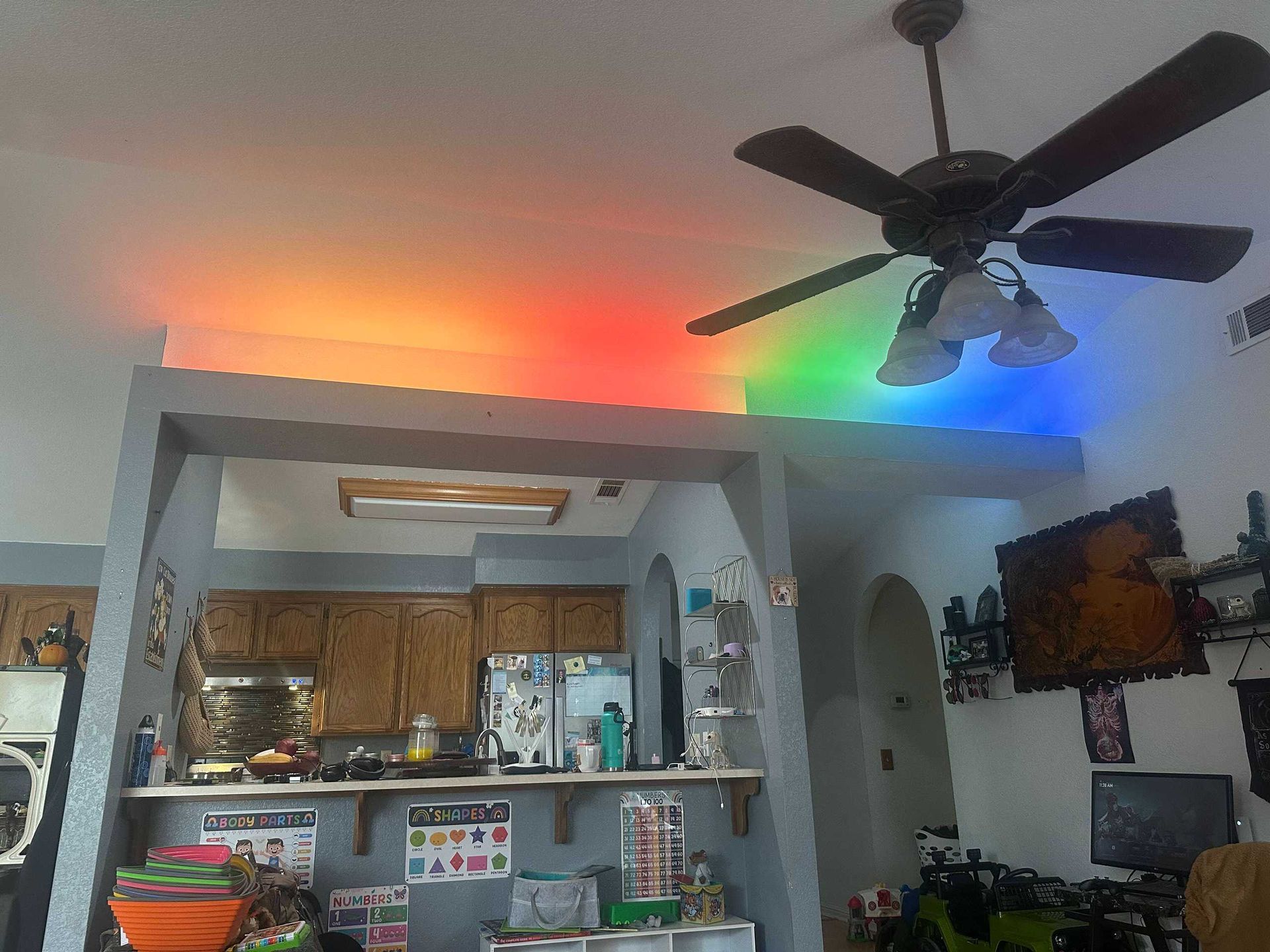 Living room with colorful ceiling lights, ceiling fan, kitchen pass-through, and wall-mounted TV