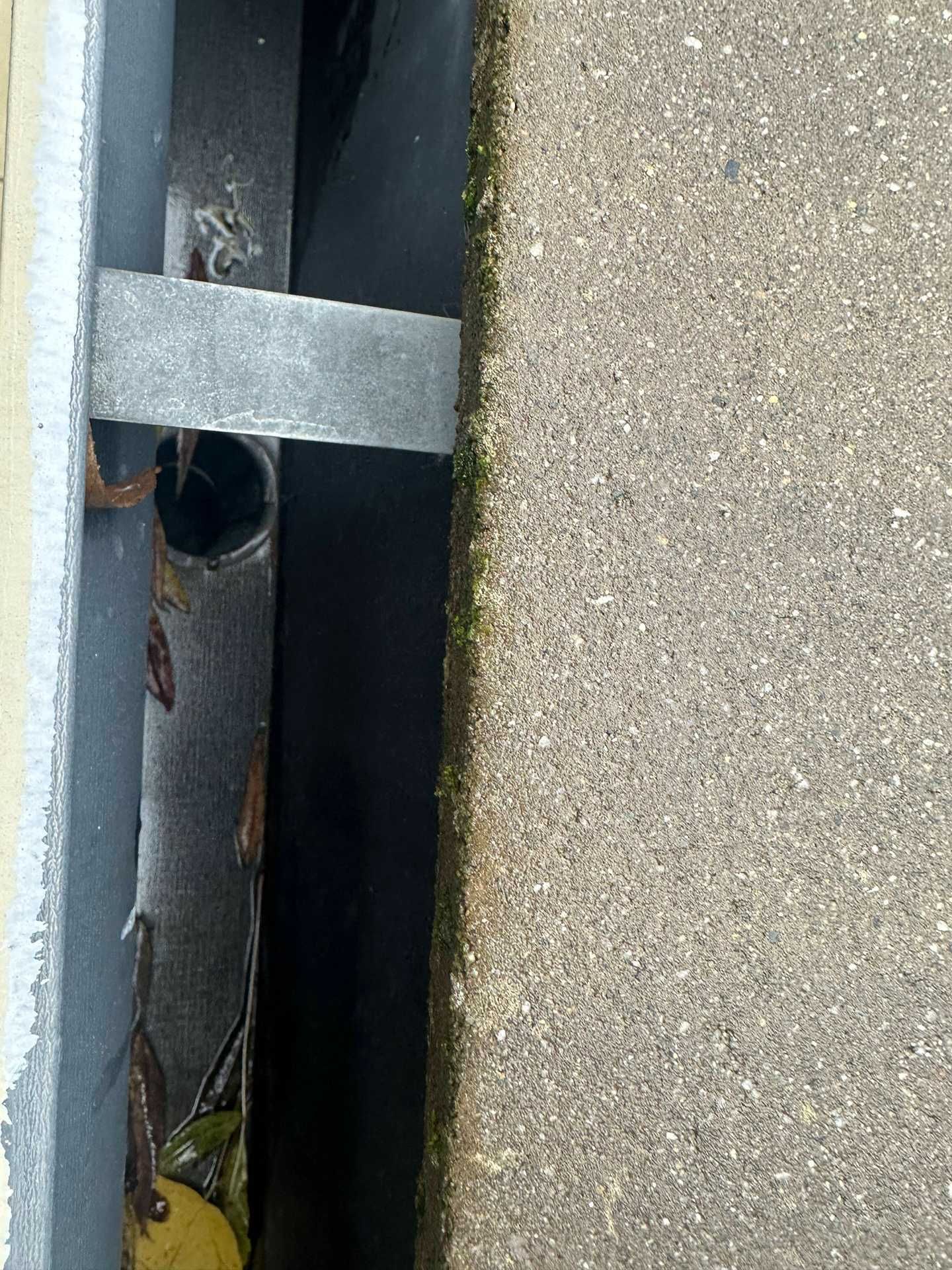 Close-up of a narrow metal drain beside gray concrete, with a dark pipe opening and debris inside.