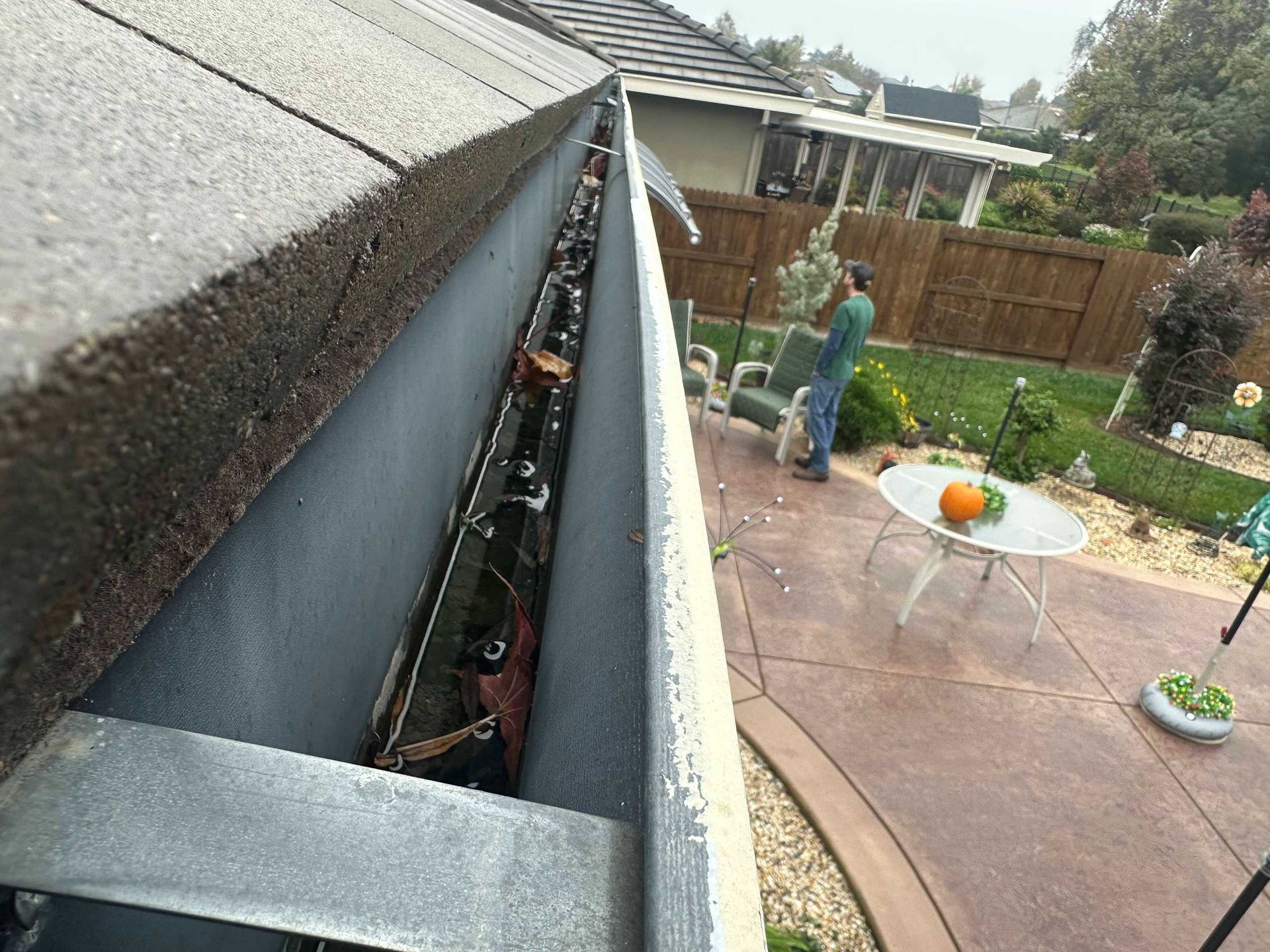 Person clearing debris from a clogged roof gutter beside a backyard patio and garden