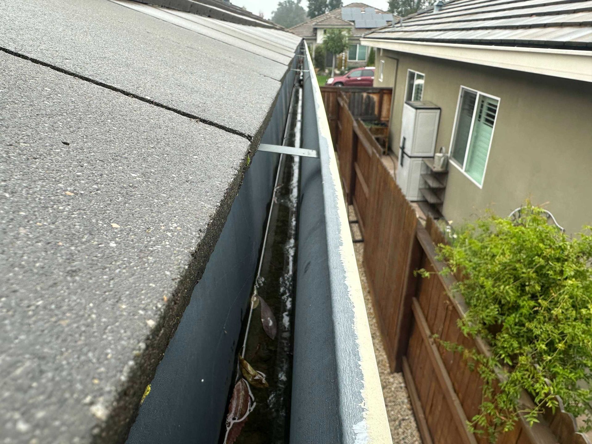 Narrow rooftop gutter gap between a gray roof and a house beside a wooden fence.
