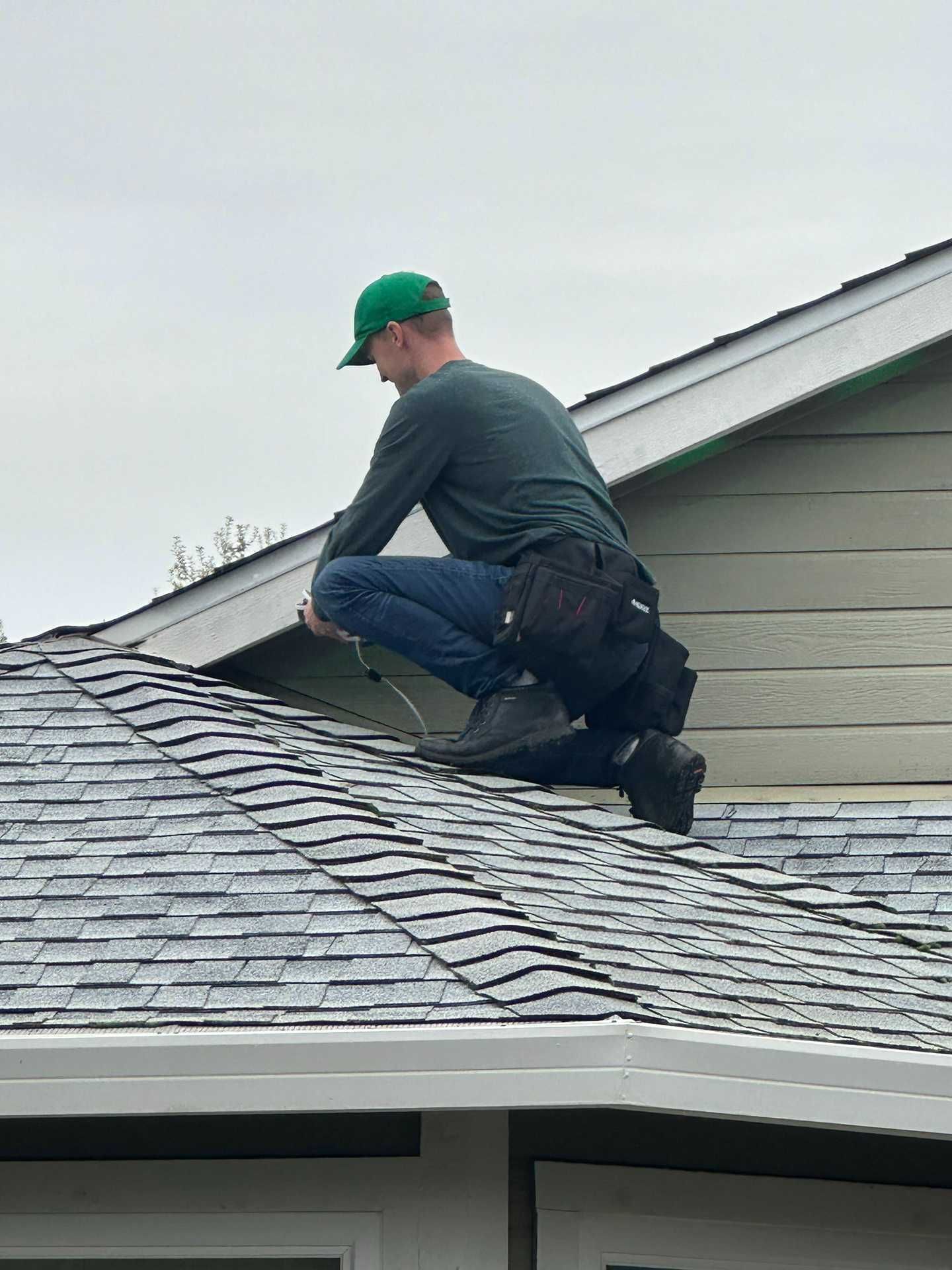Worker kneeling on a shingled roof near the ridge, inspecting or repairing it under overcast sky