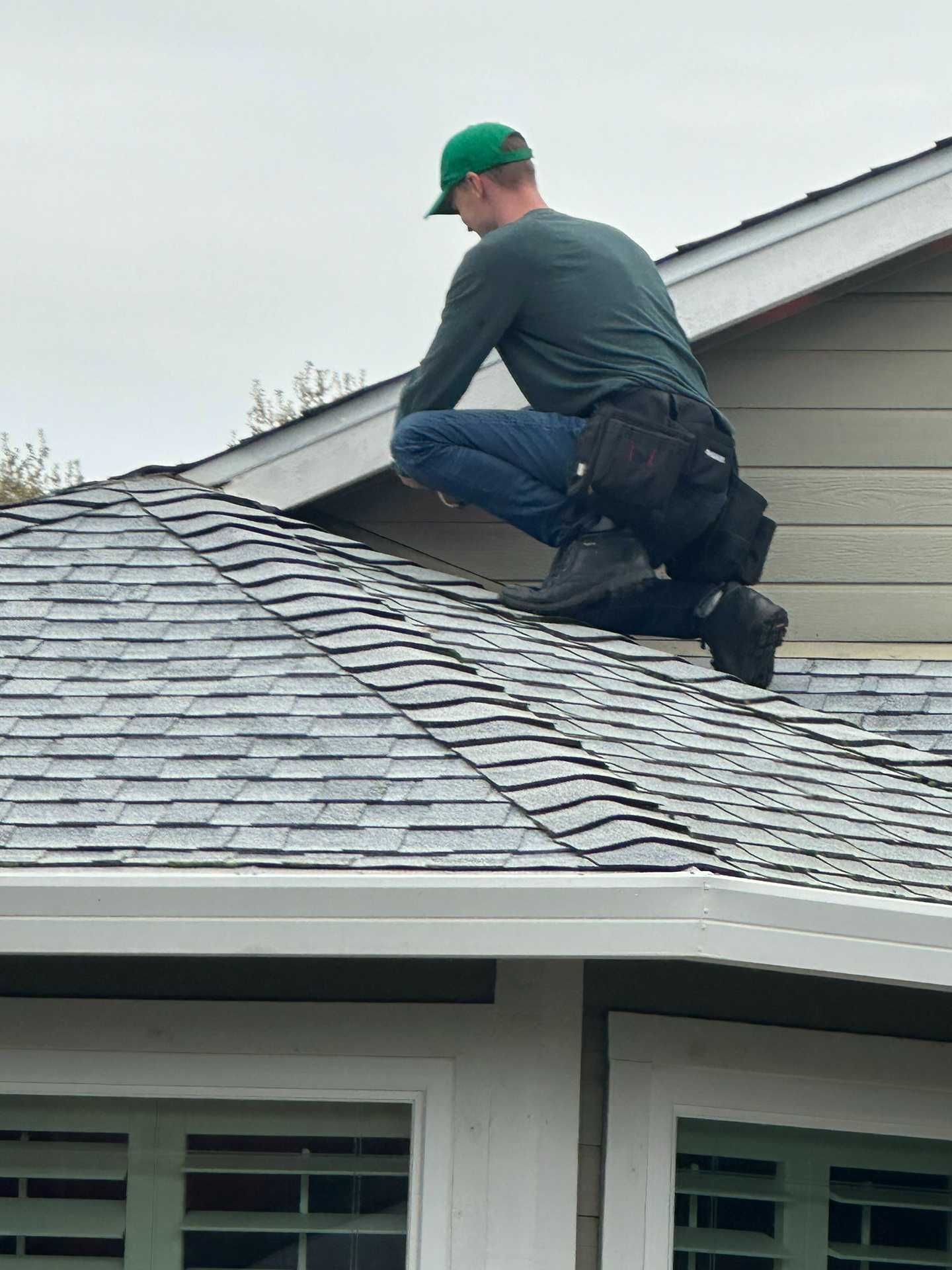 Worker kneeling on a gray shingle roof, inspecting or repairing it near a house eave.