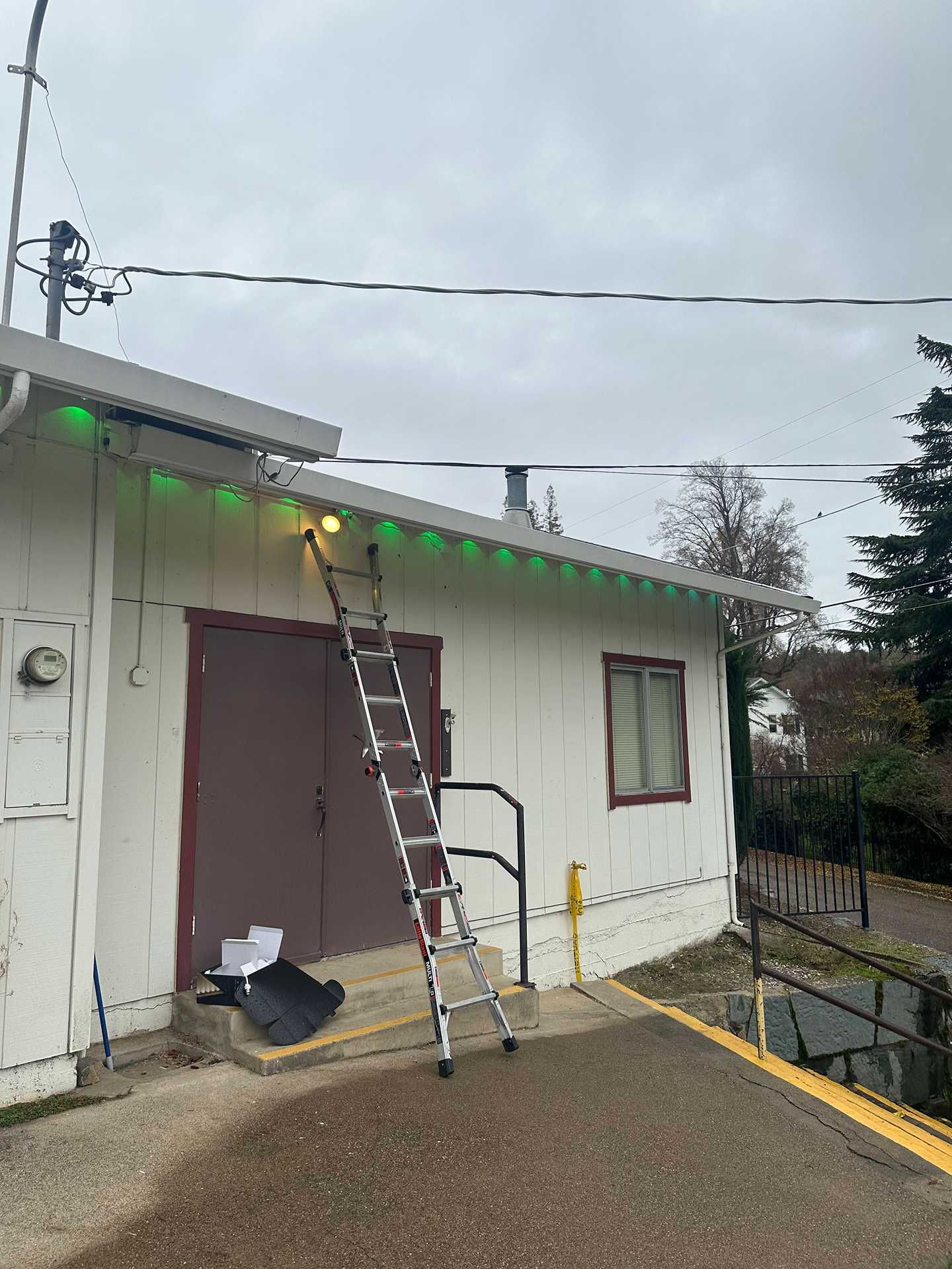 A ladder leans against a small white building with green holiday lights along the roofline.