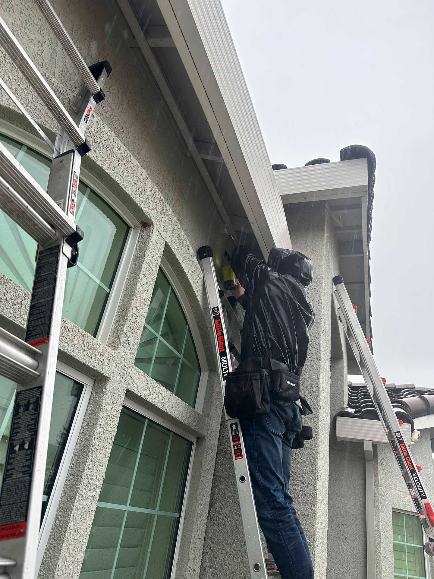 Worker on a ladder cleaning or repairing a house exterior near the roofline.