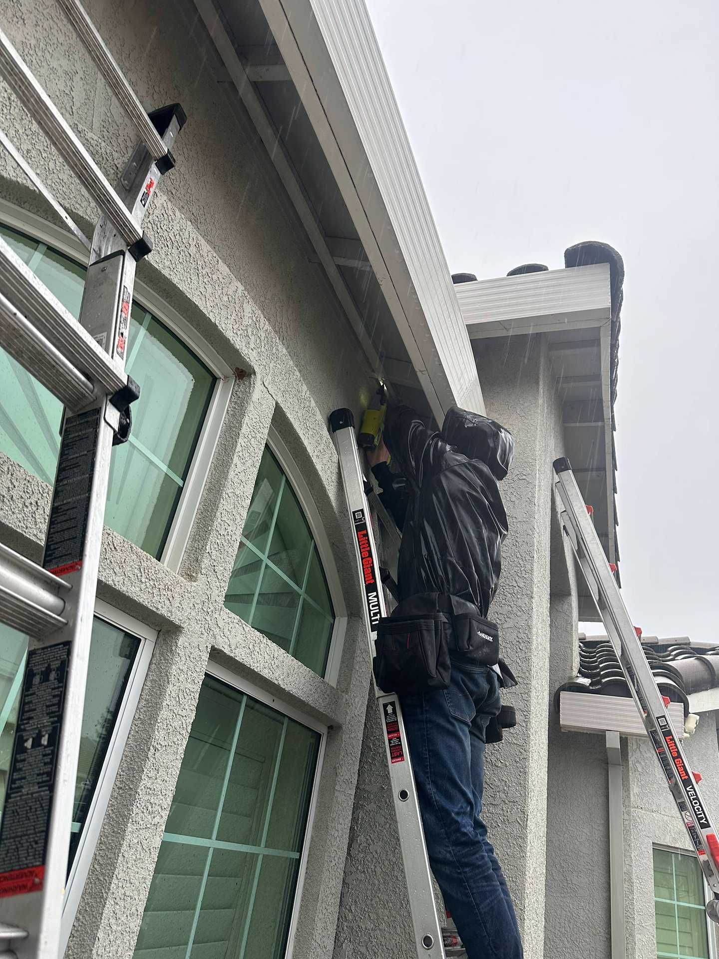 Worker on a ladder cleaning gutters beside a house with white trim and green windows.